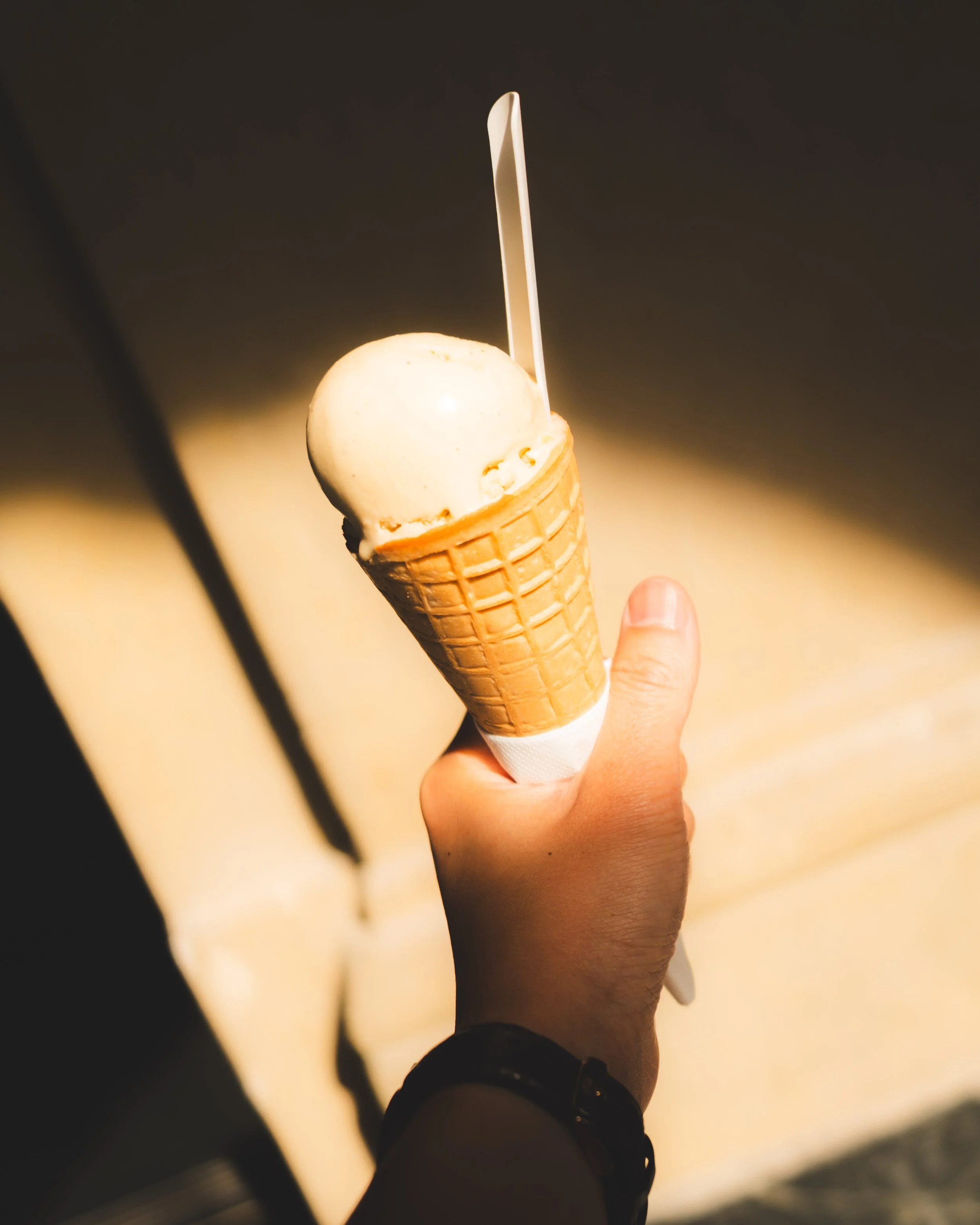 A person holding an ice cream cone with a scoop of vanilla ice cream and a plastic spoon in it, against a blurred background.