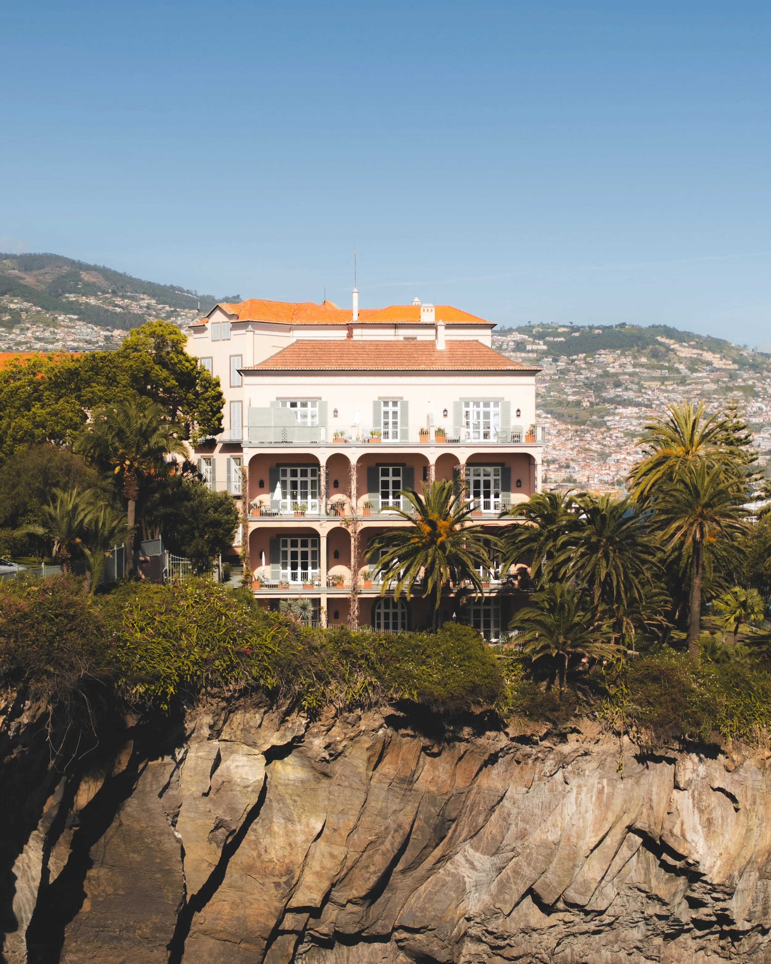 A view of a the Reid's Palace hotel with orange-tile roof and white walls, partly obscured by lush green trees, with a hillside cityscape in the background under a clear blue sky. The luxury hotel is located above a rocky cliff.