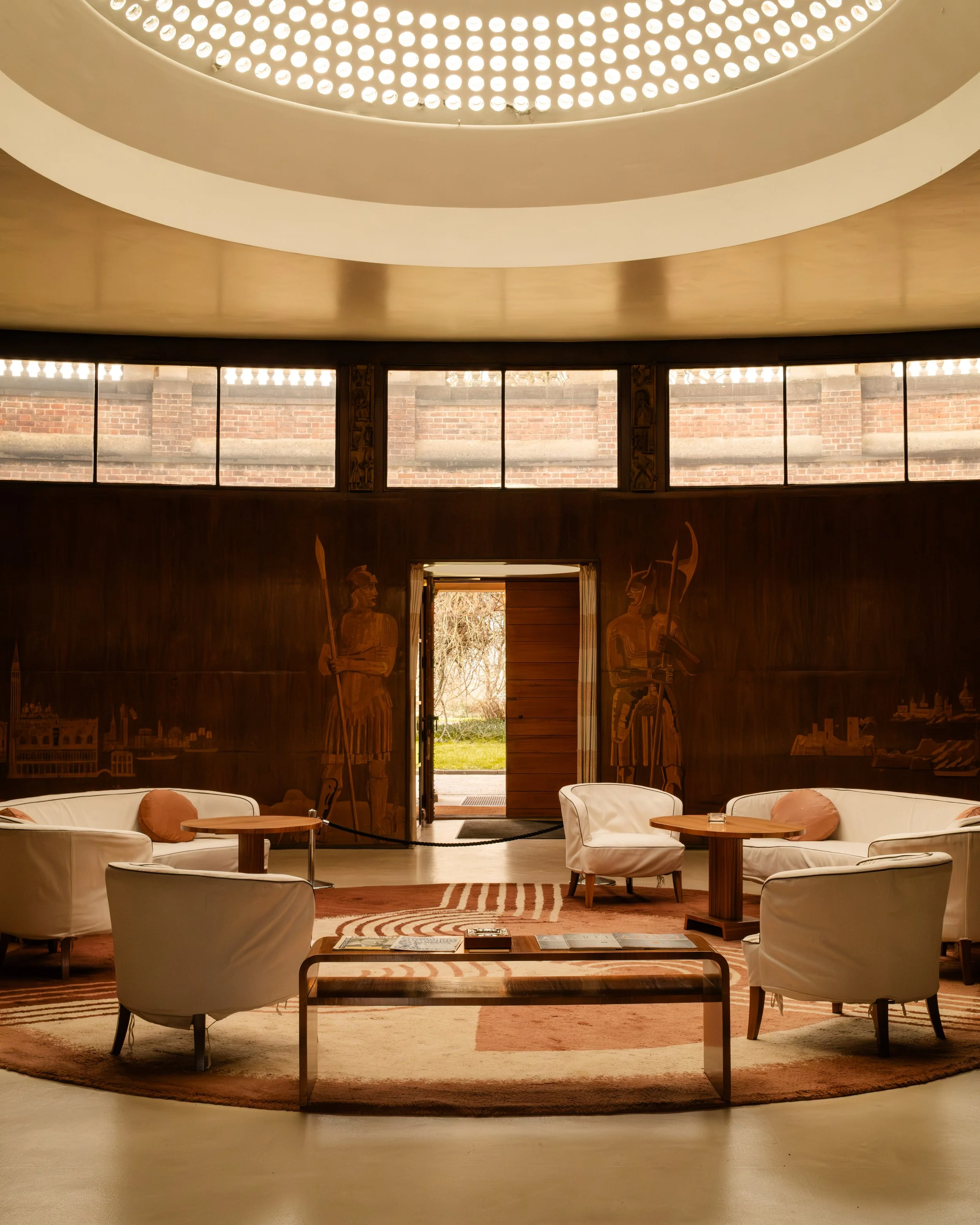 Interior of a round room with wooden walls, featuring two large carved statues of armored warriors, a circular arrangement of white chairs and armchairs, a wooden table, and a chandelier on the ceiling. Lionel Chu - Hotel & Travel Photographer