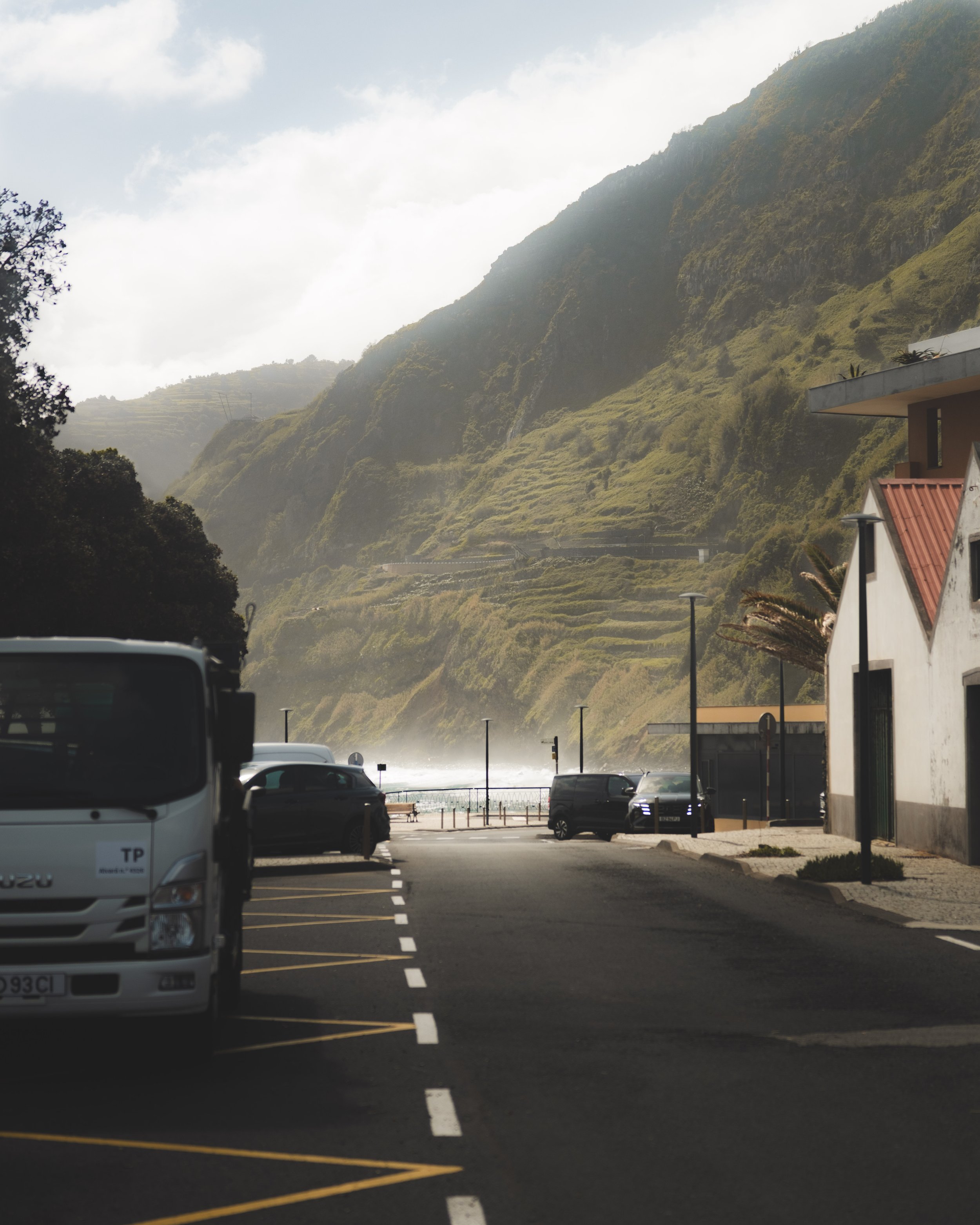 A street on a sunny day with a mountain in the background, several parked cars, and a few buildings and street lamps in the foreground.