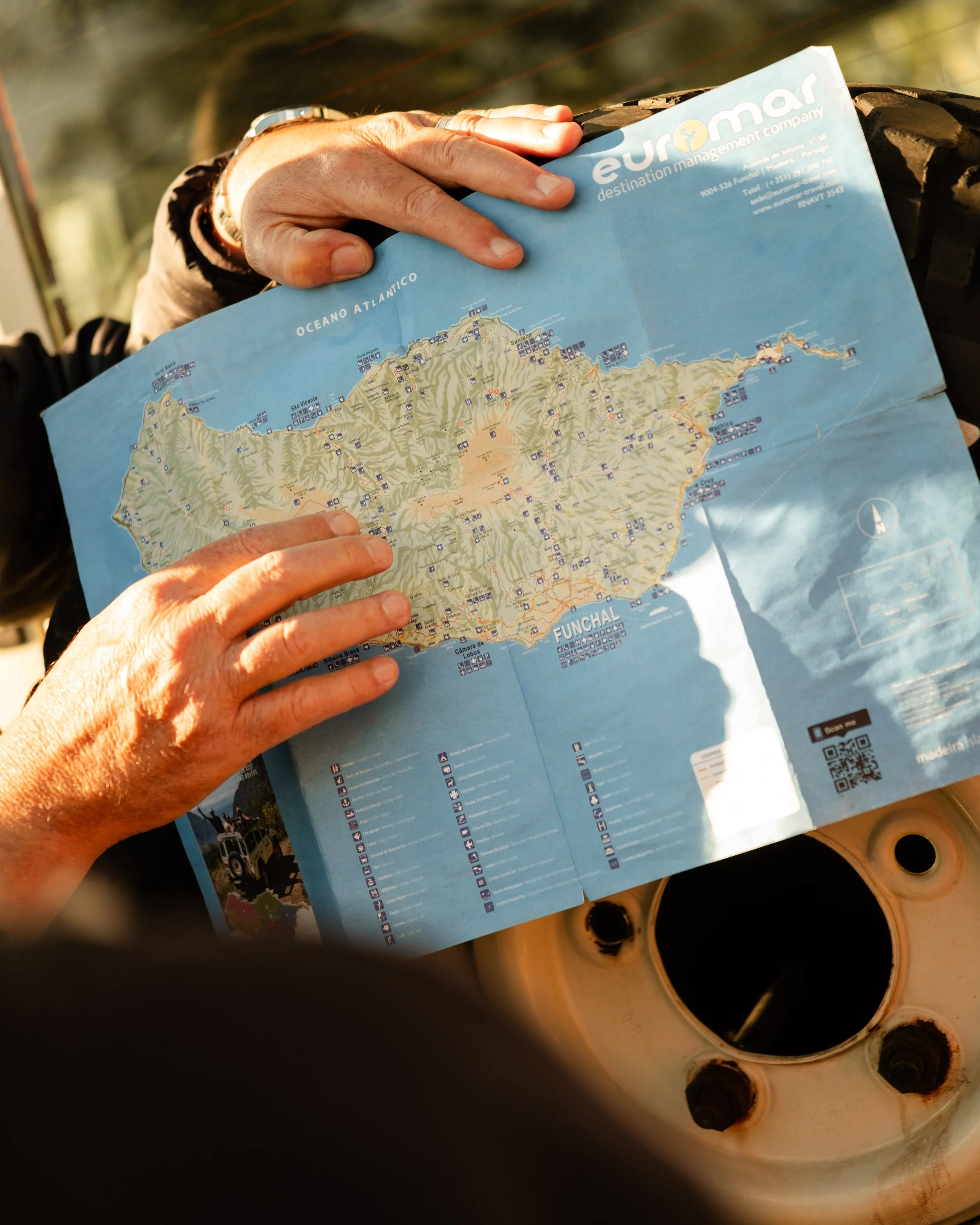 A person is holding a blue map of the Funchal area in Portugal, with a hand on the coast near the Atlantic Ocean, standing over a circular, rusted car wheel rim.