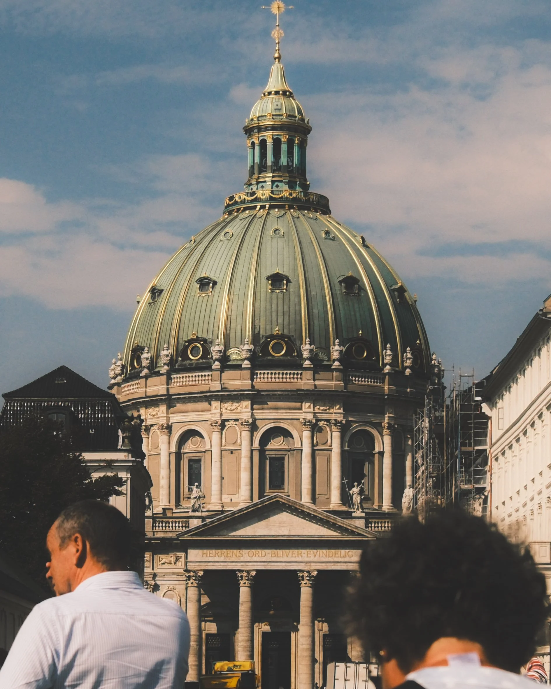 The image shows the dome of a historic building with intricate architectural details, a golden spire on top, and a surrounding clear blue sky. In the foreground, several people are partially visible, suggesting a public or tourist setting.