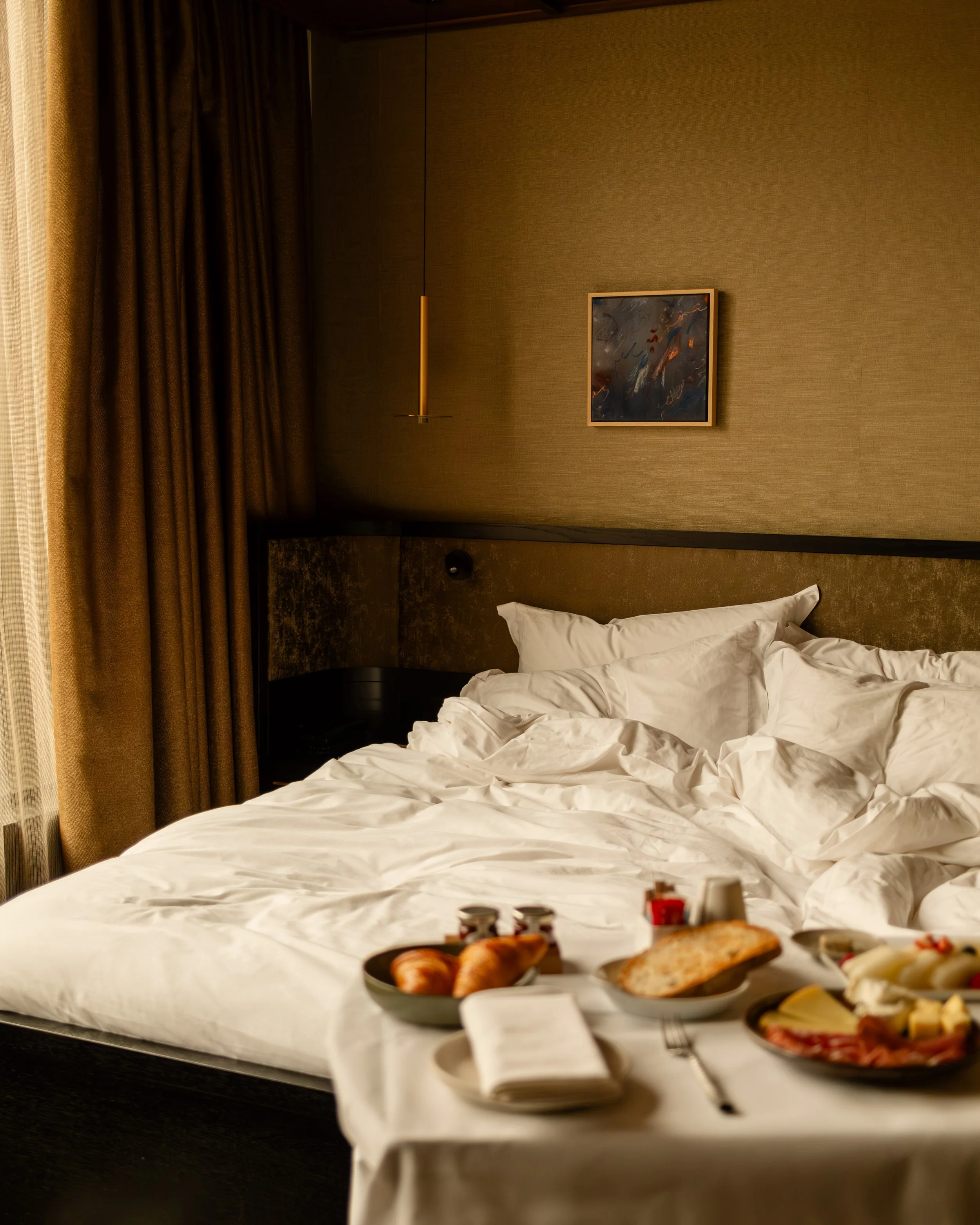 Luxury hotel room with unmade bed and breakfast tray featuring croissants, bread, cheese, and coffee. Lionel Chu - Hotel & Travel Photographer