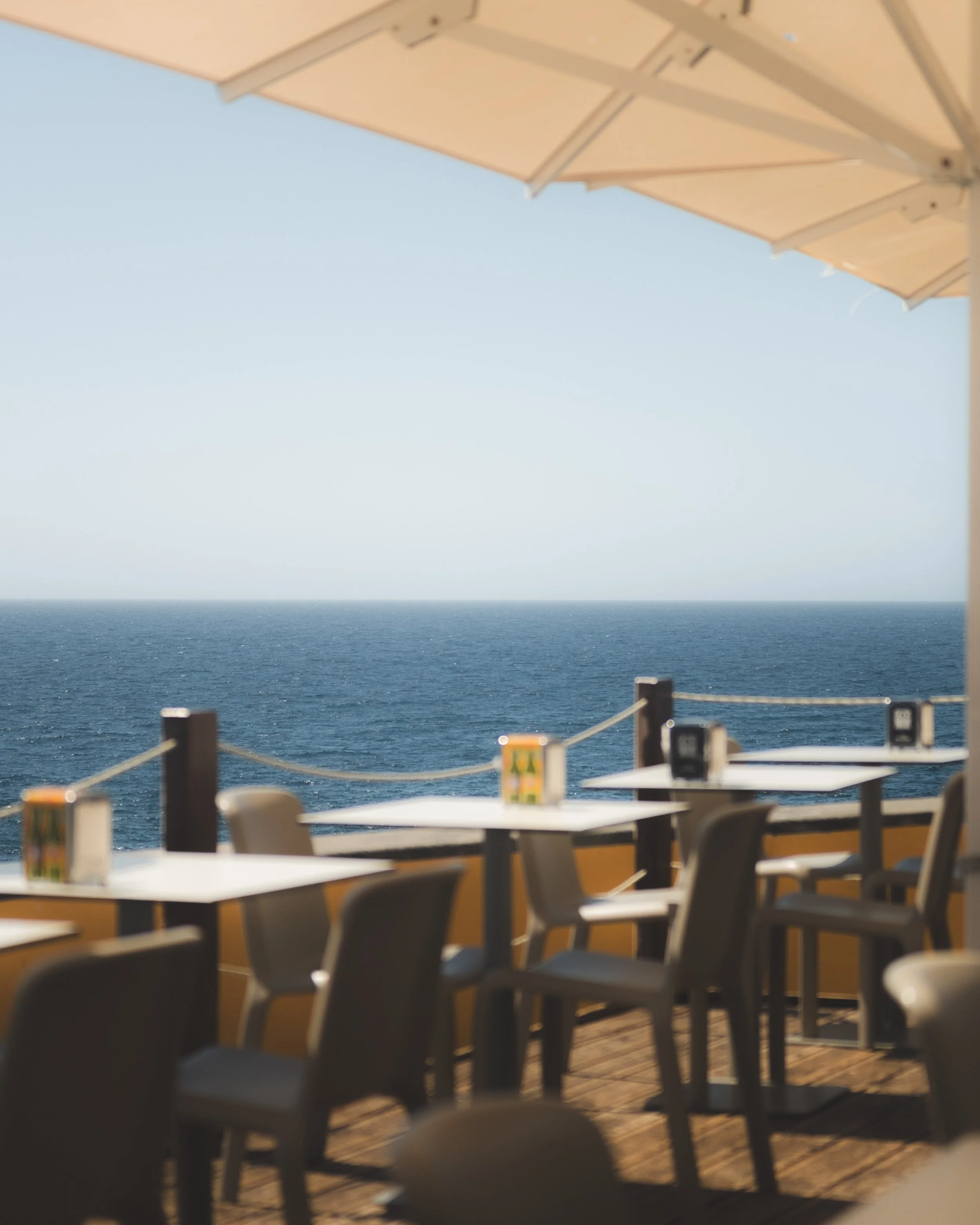 Blank tables and chairs outside on a deck or patio overlooking the ocean with a railing. The sky is clear and sunny.