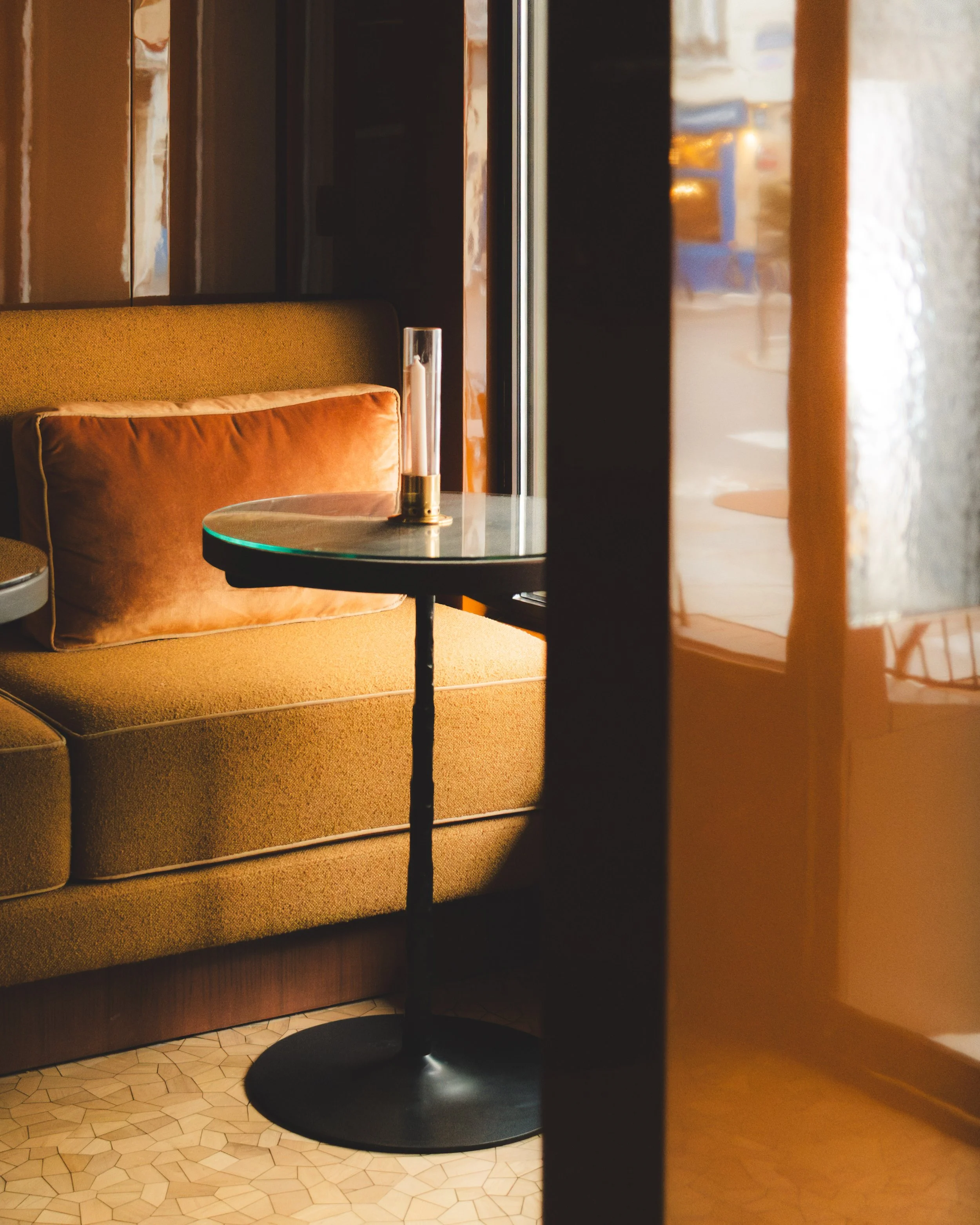 A view of a cozy seating area with an orange sofa, a round glass table, and a small decorative object, seen through a partially open door in a warmly lit room.