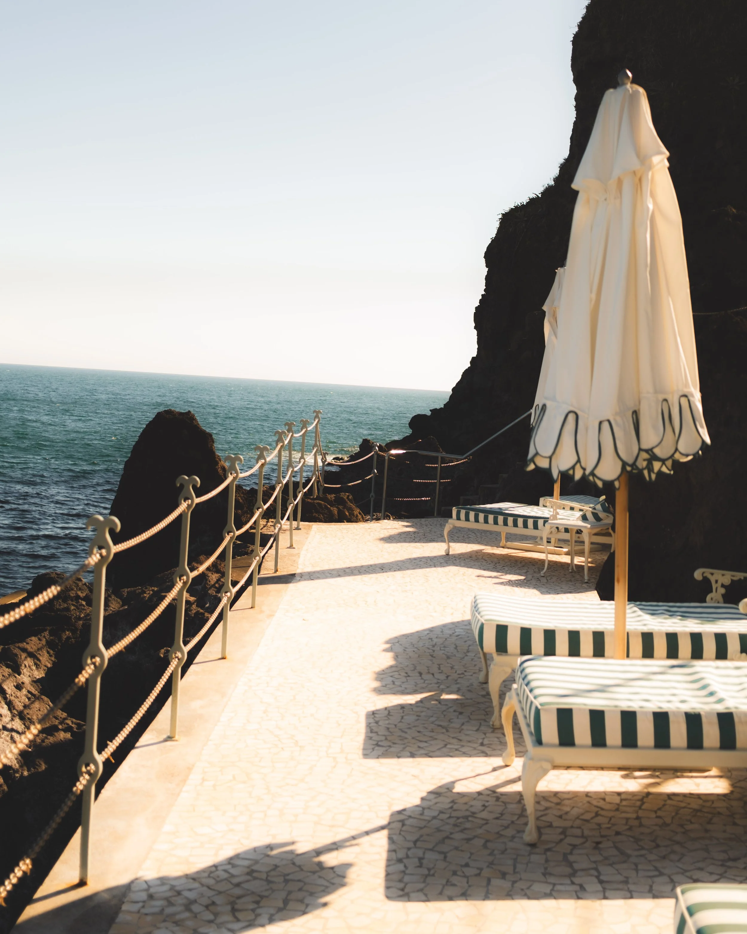 Beachside patio with striped lounge chairs, a large white umbrella, a stone pathway, black rock formations, a railing with rope, ocean view, and cliffs in the background.