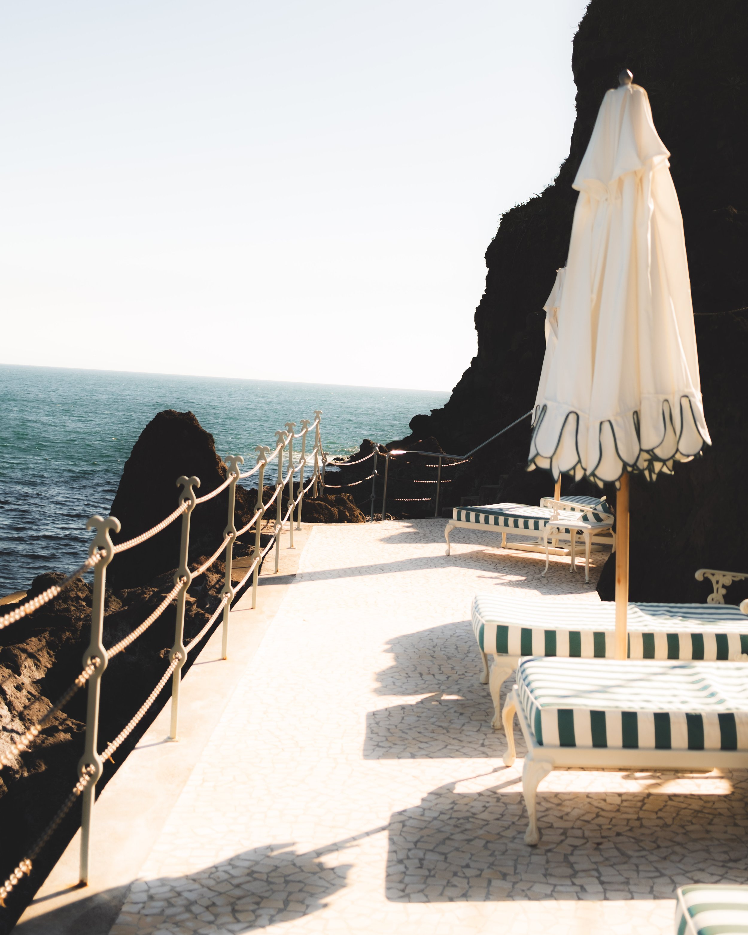 A view of a seaside patio with striped lounge chairs and a white umbrella on the right, overlooking the ocean with rocky outcrops and a hazy sky in the background.