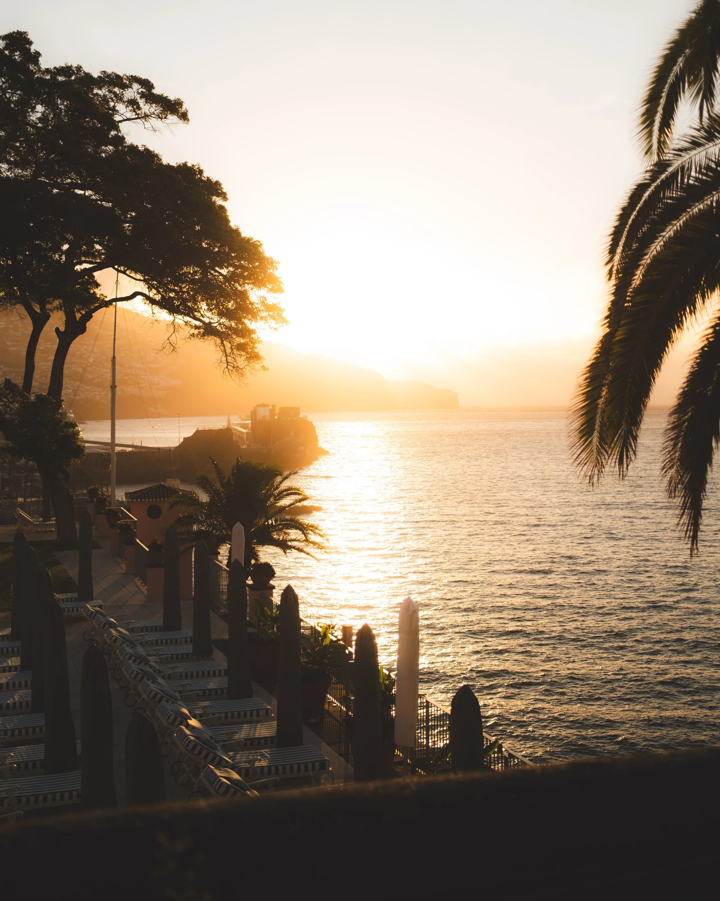 Sunset over a body of water with trees on the shoreline, and a row of lounge chairs with striped cushions in the foreground. Picture from a luxury hotel. Lionel Chu - Hotel & Travel Photographer