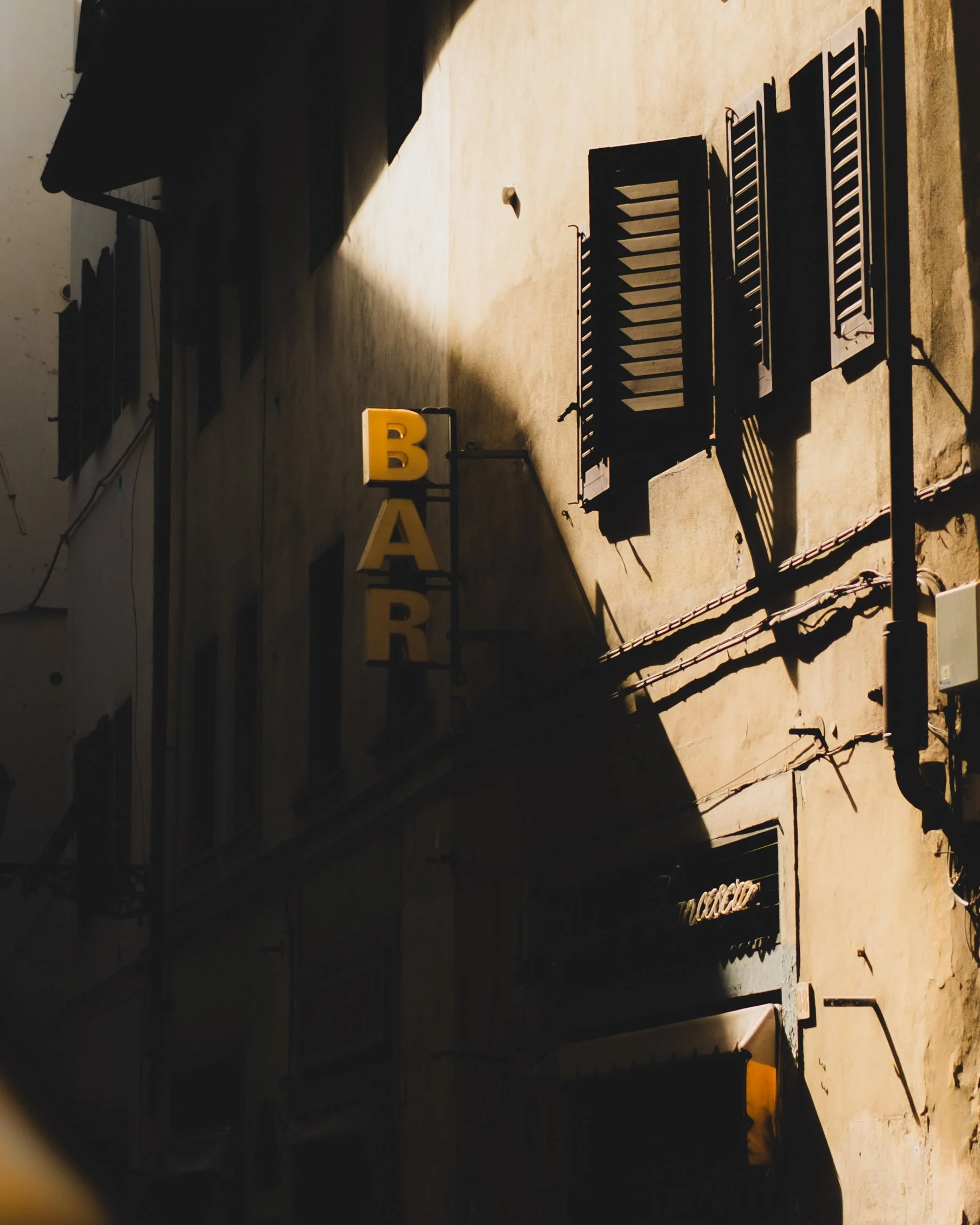 Sunlit beige building with black shutters, a vertical yellow sign spelling 'BAR', and a black sign with white cursive text. Lionel Chu - Hotel & Travel Photographer