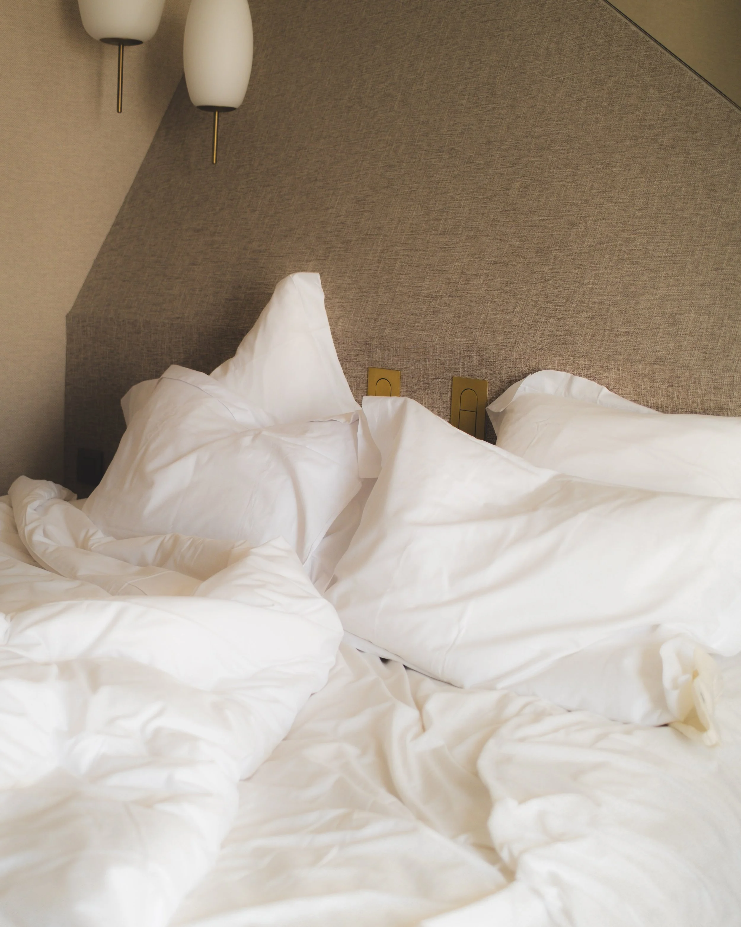 The image shows a bed with white rumpled sheets and pillows in front of a textured beige headboard. Two lamps with white shades and gold accents are mounted on the wall above the headboard. There are also two electrical outlets visible on the headboard.