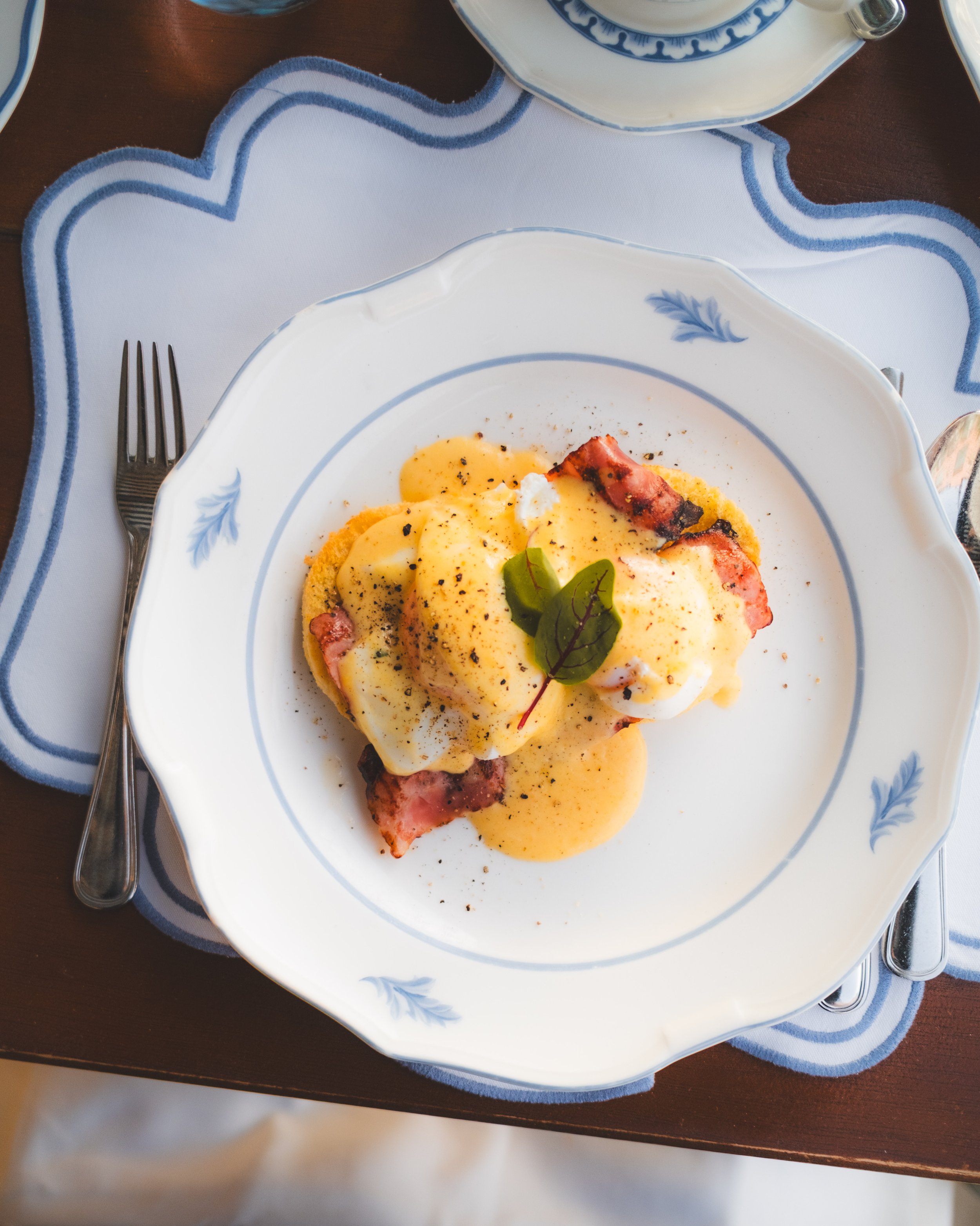 Plate of eggs Benedict with hollandaise sauce, bacon, and garnished with greens on a white and blue decorative plate, placed on a white tablecloth with blue trim.
