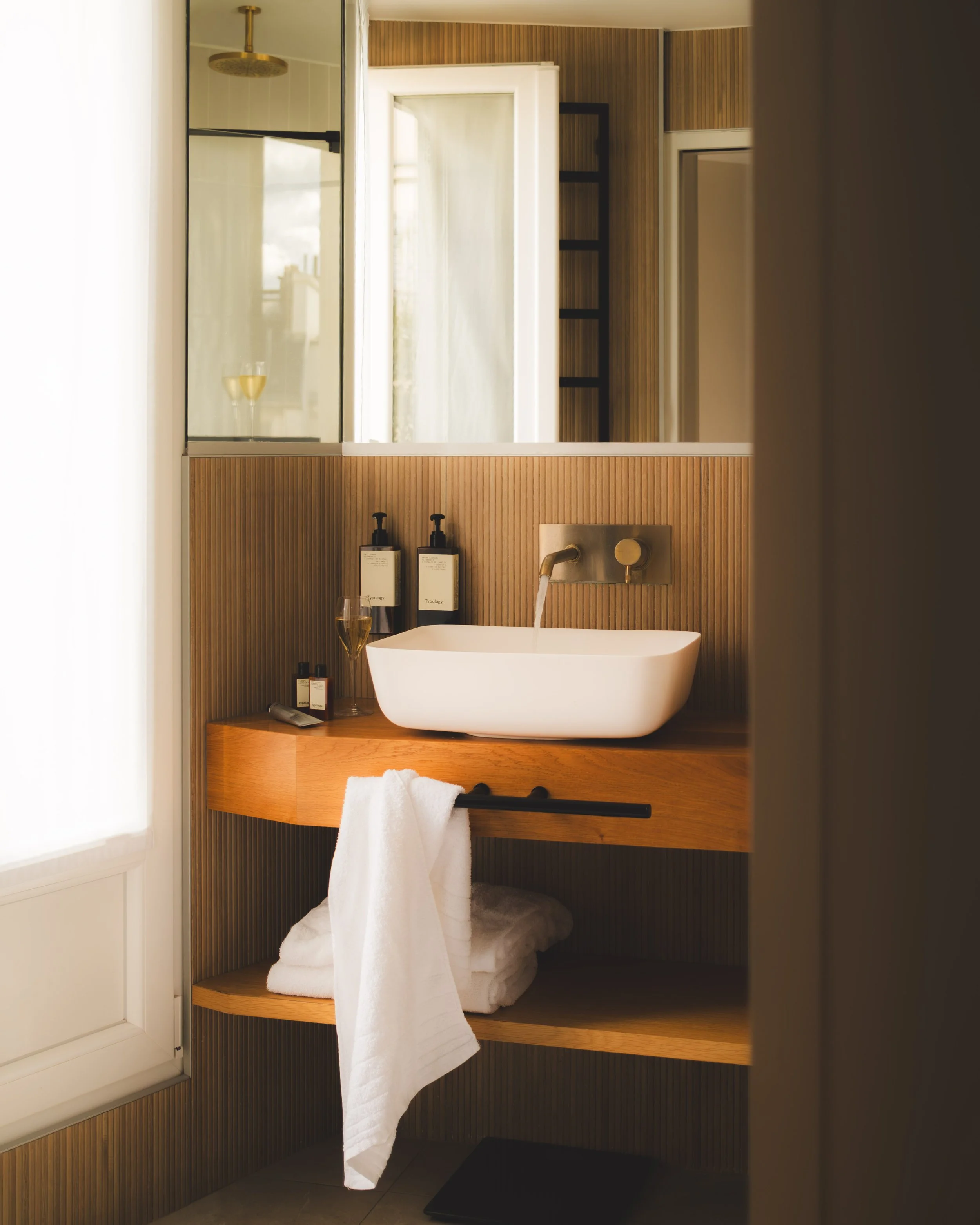 A bathroom with a white bathtub, wood accents, a large mirror, black shelf, window, towel bar with white towel, soap dispensers, and a glass of wine.