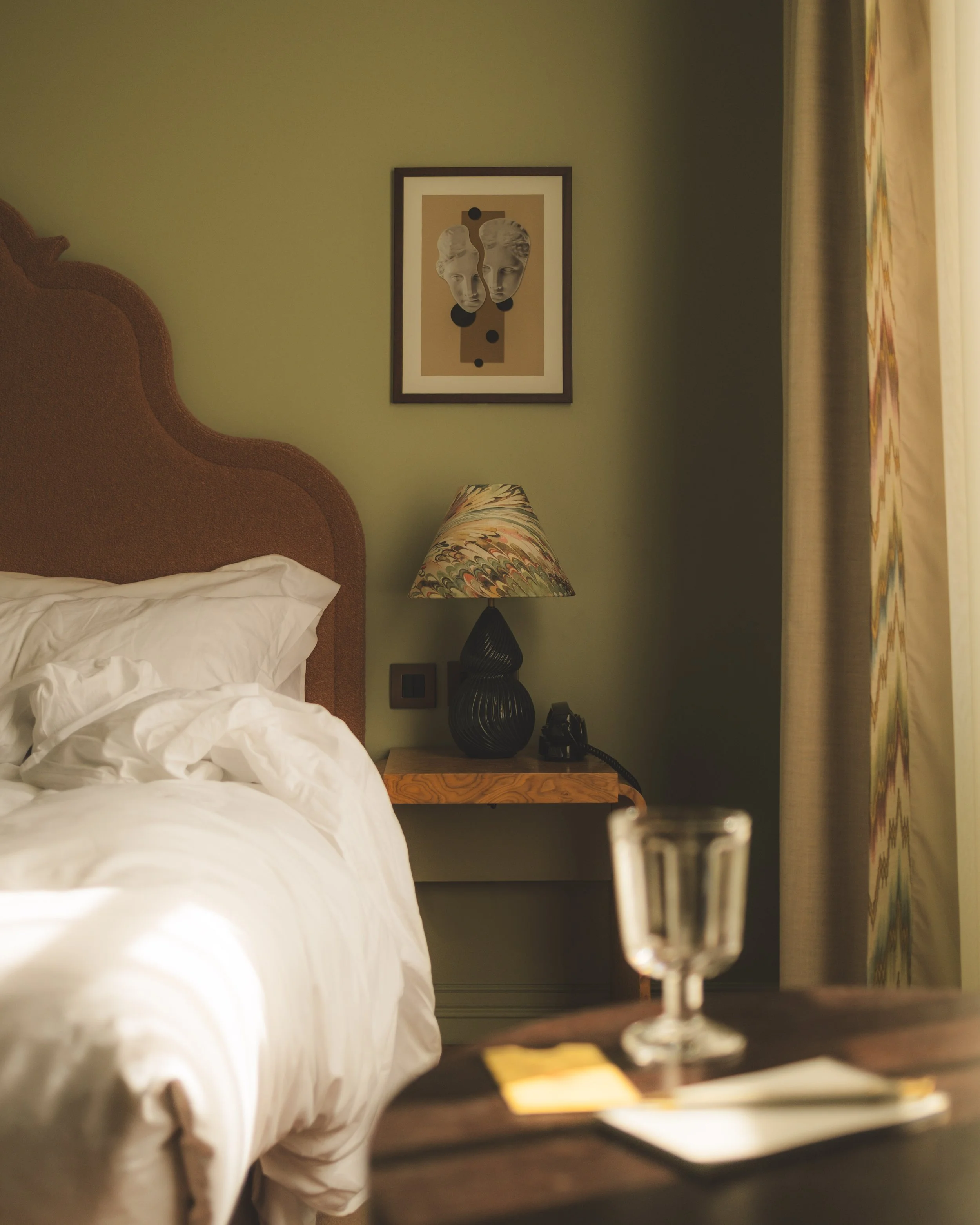 A bedroom with a bed on the left and a nightstand on the right, featuring a black table lamp with a colorful shade, and a framed art piece with classical faces on the green wall behind.