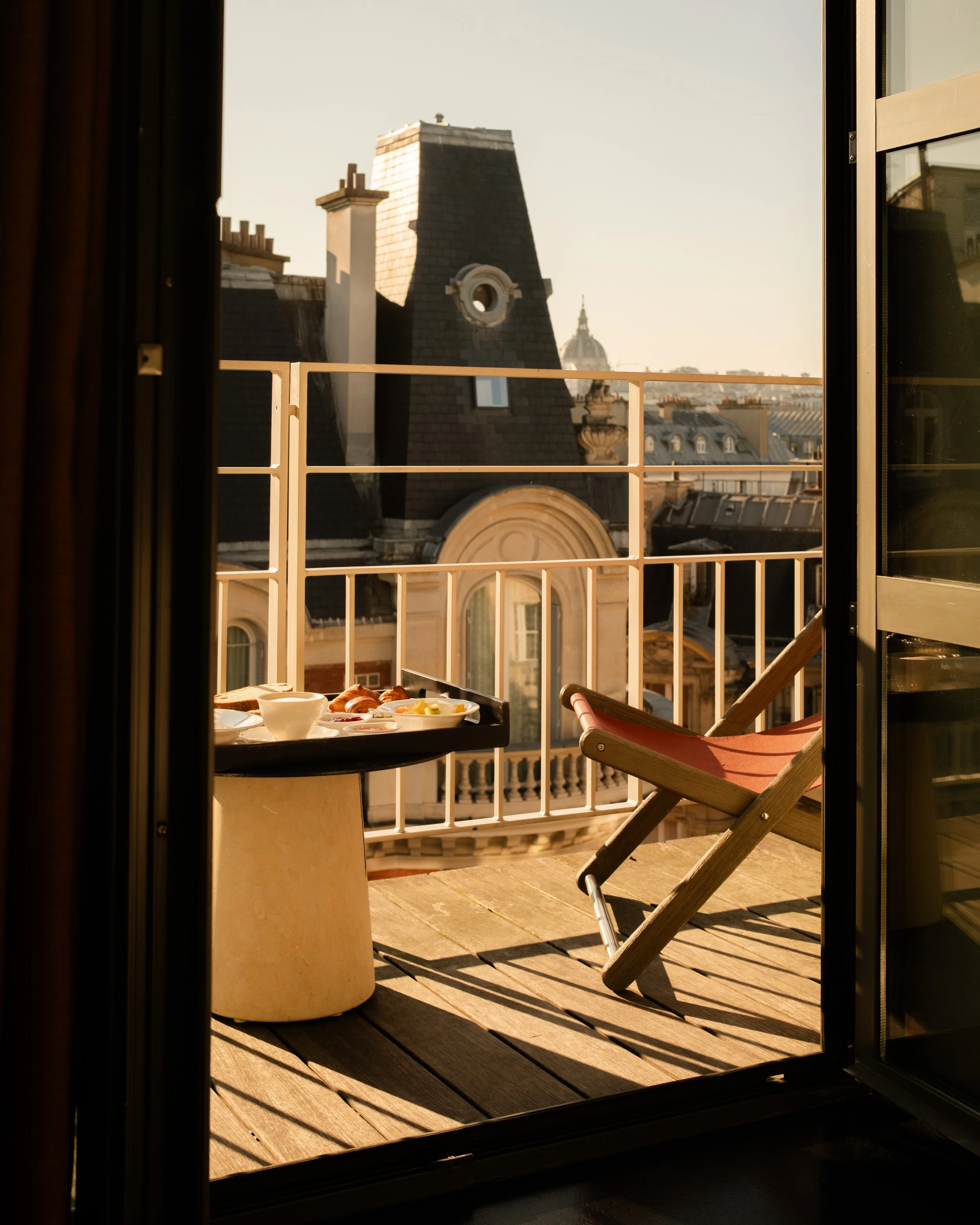 A balcony with a reclining chair and a small table set with breakfast, overlooking Paris rooftops and buildings, in the early morning sunlight. Lionel Chu - Hotel & Travel Photographer