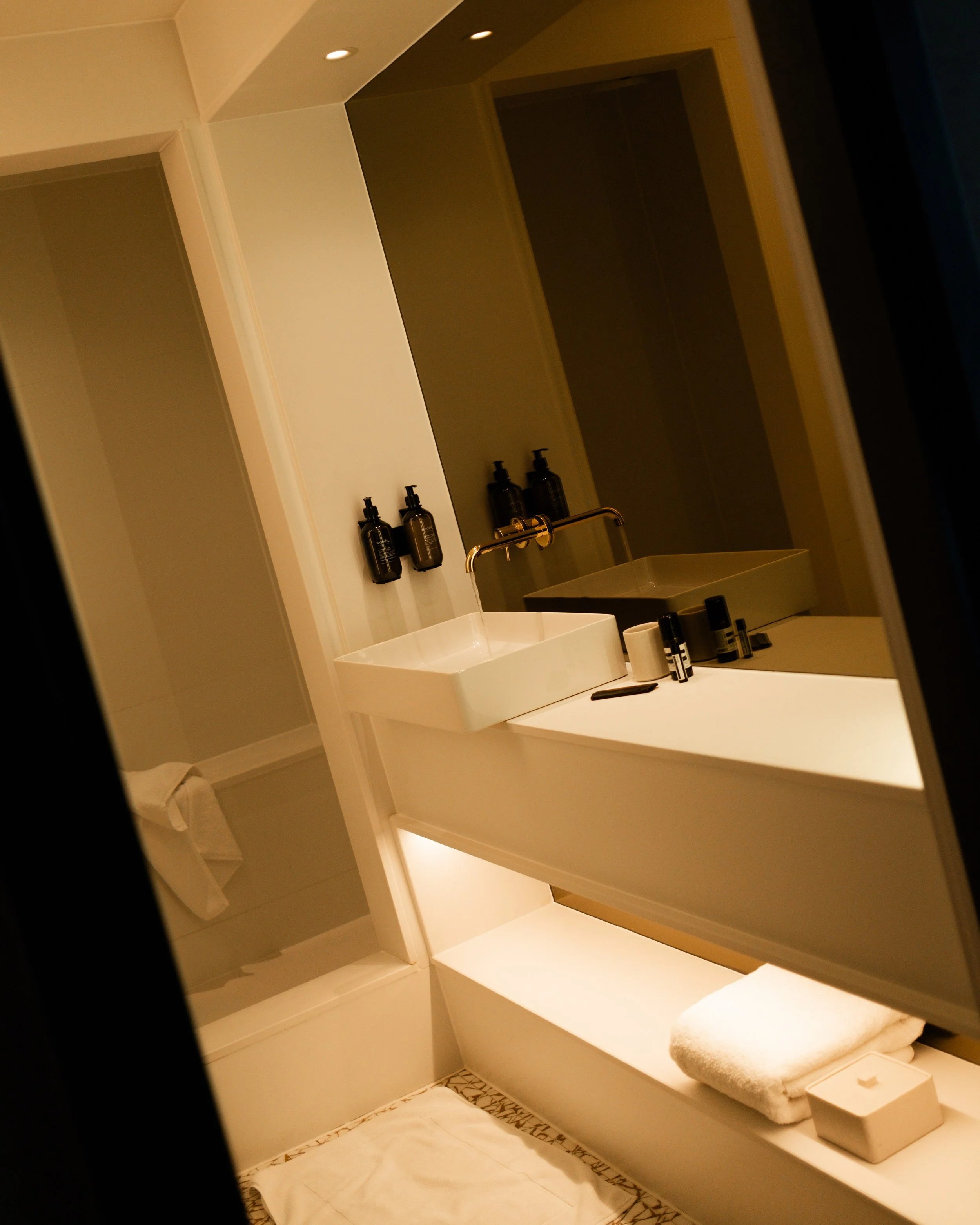 Modern bathroom with white sink, black wall-mounted soap dispensers, mirror, and lit shelf with towel and toiletries. Lionel Chu - Hotel & Travel Photographer