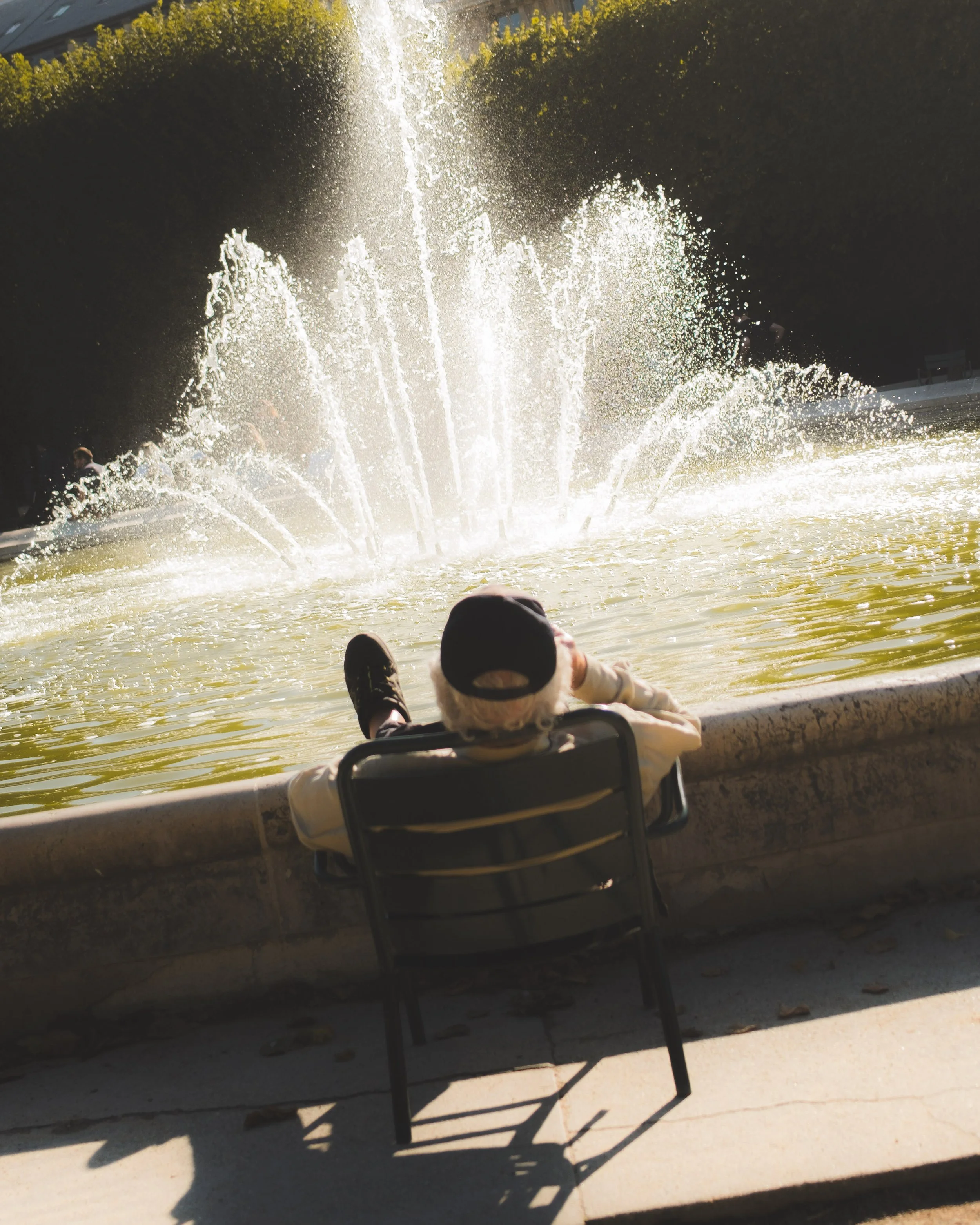 A person with a hat lying on a chair by a fountain with water spraying upwards, shaded by trees and buildings in the background.