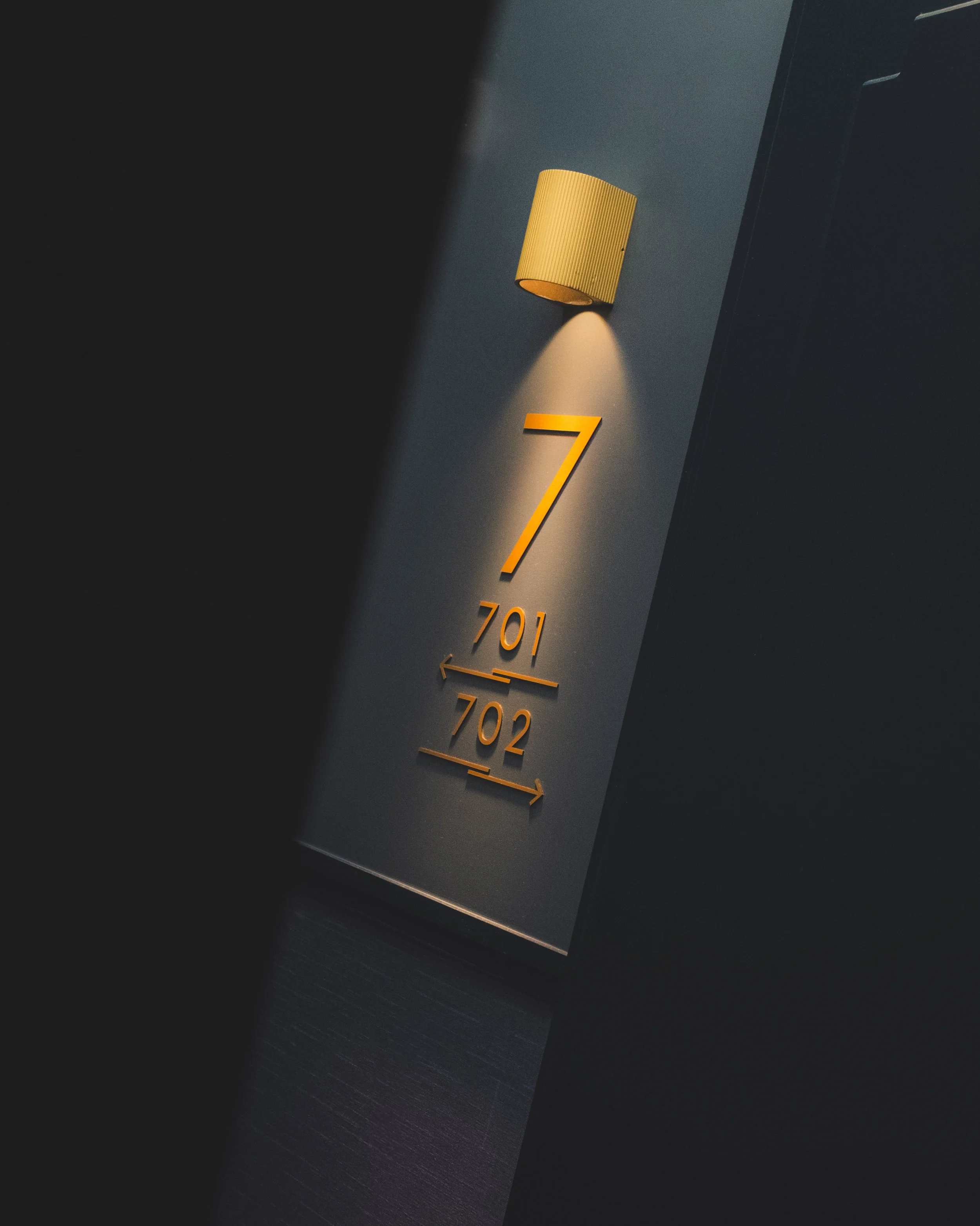 Close-up of a wall-mounted yellowish wall sconce light fixture illuminating a numbered hotel room sign with the number 701 and slightly below a 702. The photo is taken at an angle in low light conditions, with the background dark.