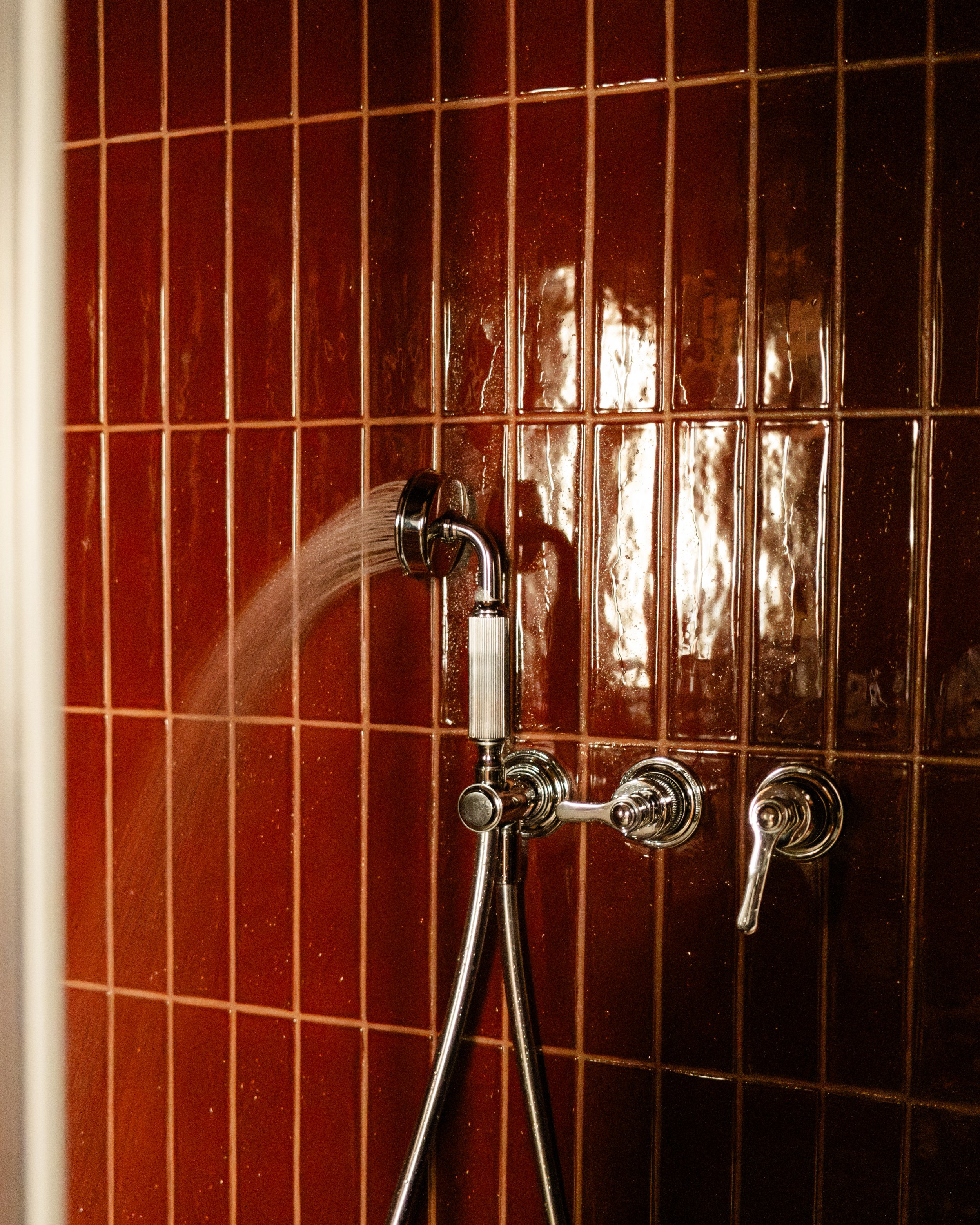Close-up of a showerhead and faucet with a red tiled wall in the background. Lionel Chu - Hotel & Travel Photography