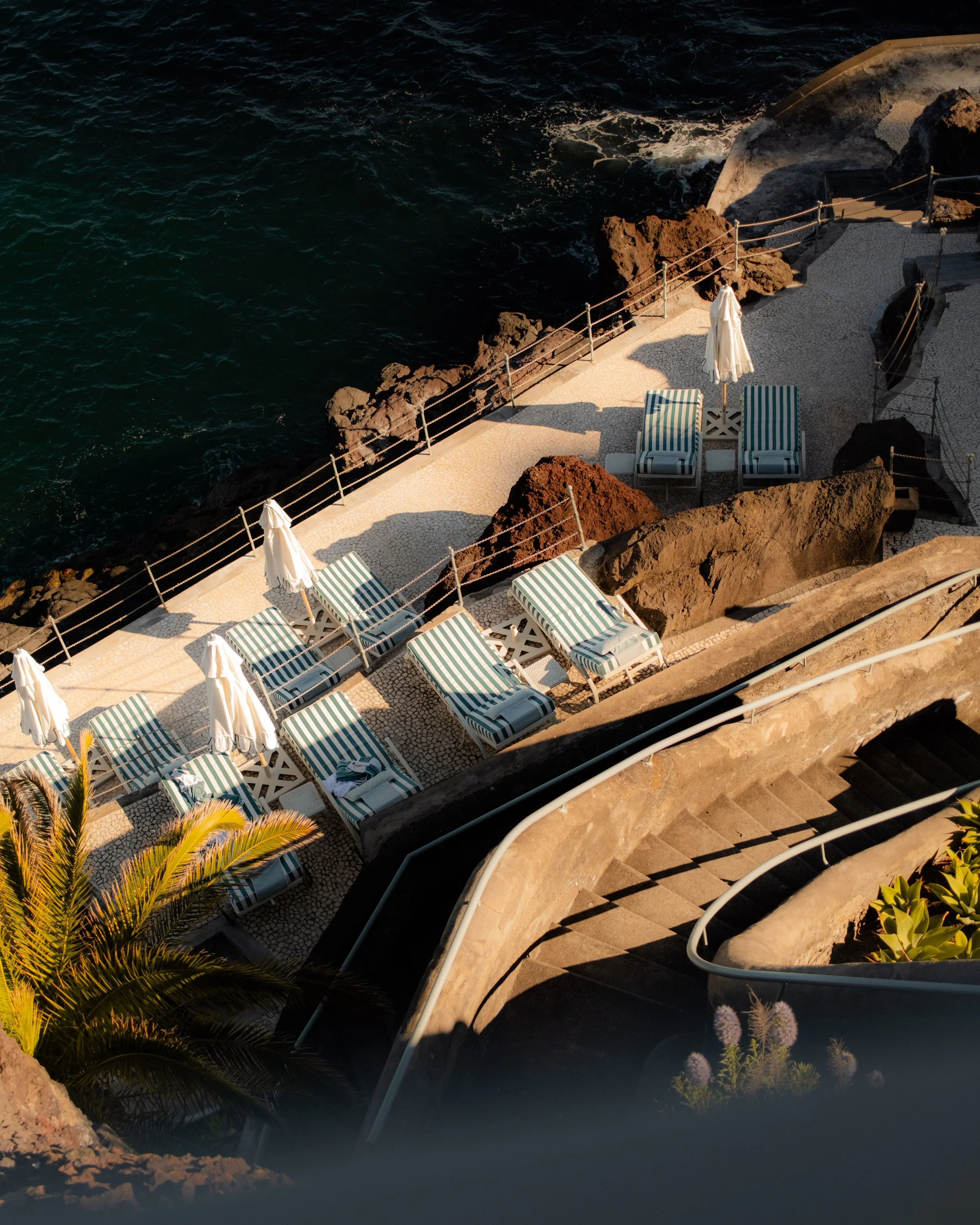 A seaside scene showing a rocky coastline with a paved sunbathing area with striped lounge chairs, umbrellas, and a staircase leading to the water, with dark blue ocean waves in the background.
