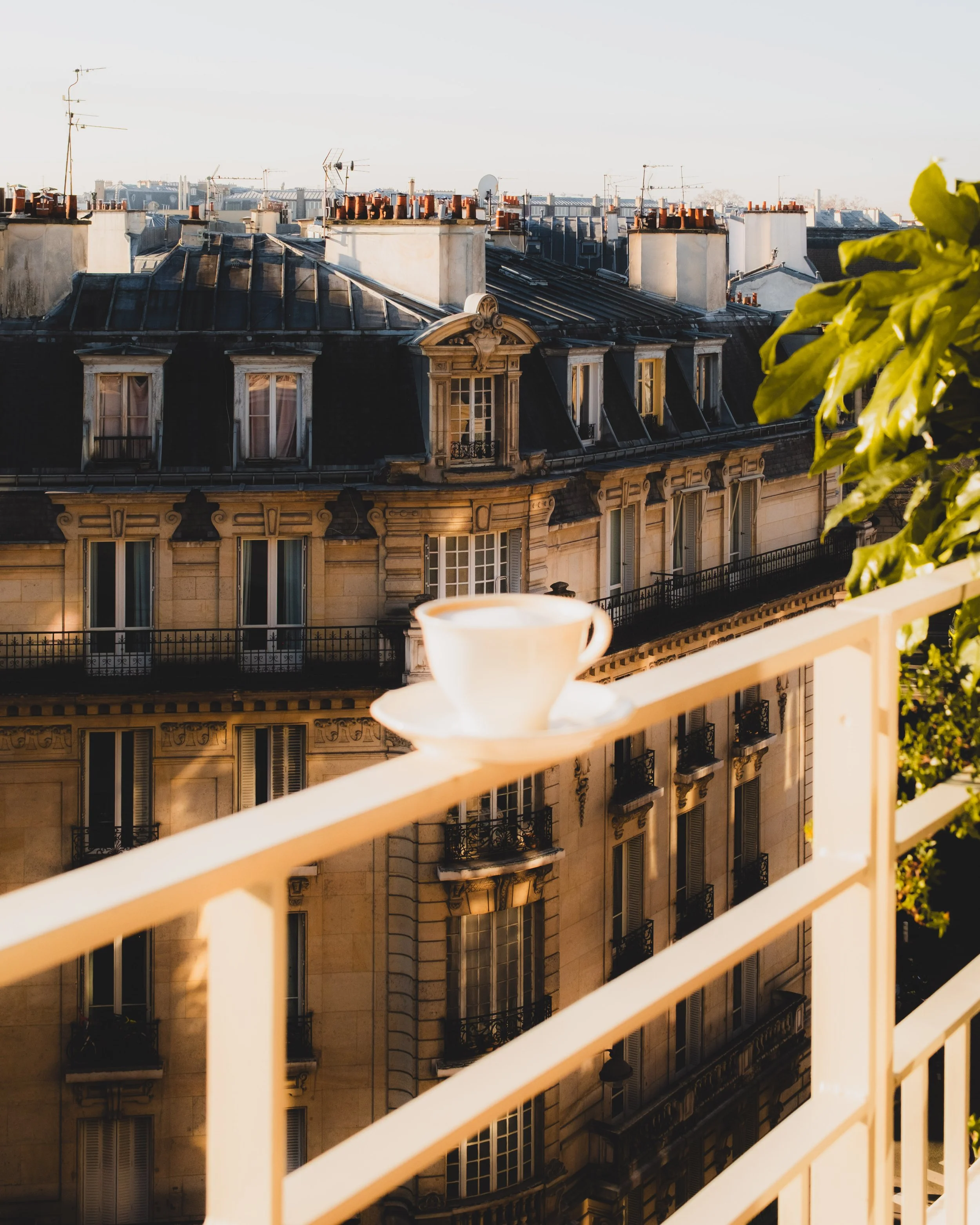 View of Paris rooftops with chimneys, windows, and a light-colored building facade, seen from a balcony with a white railing and a coffee cup in the foreground, during daylight hours.