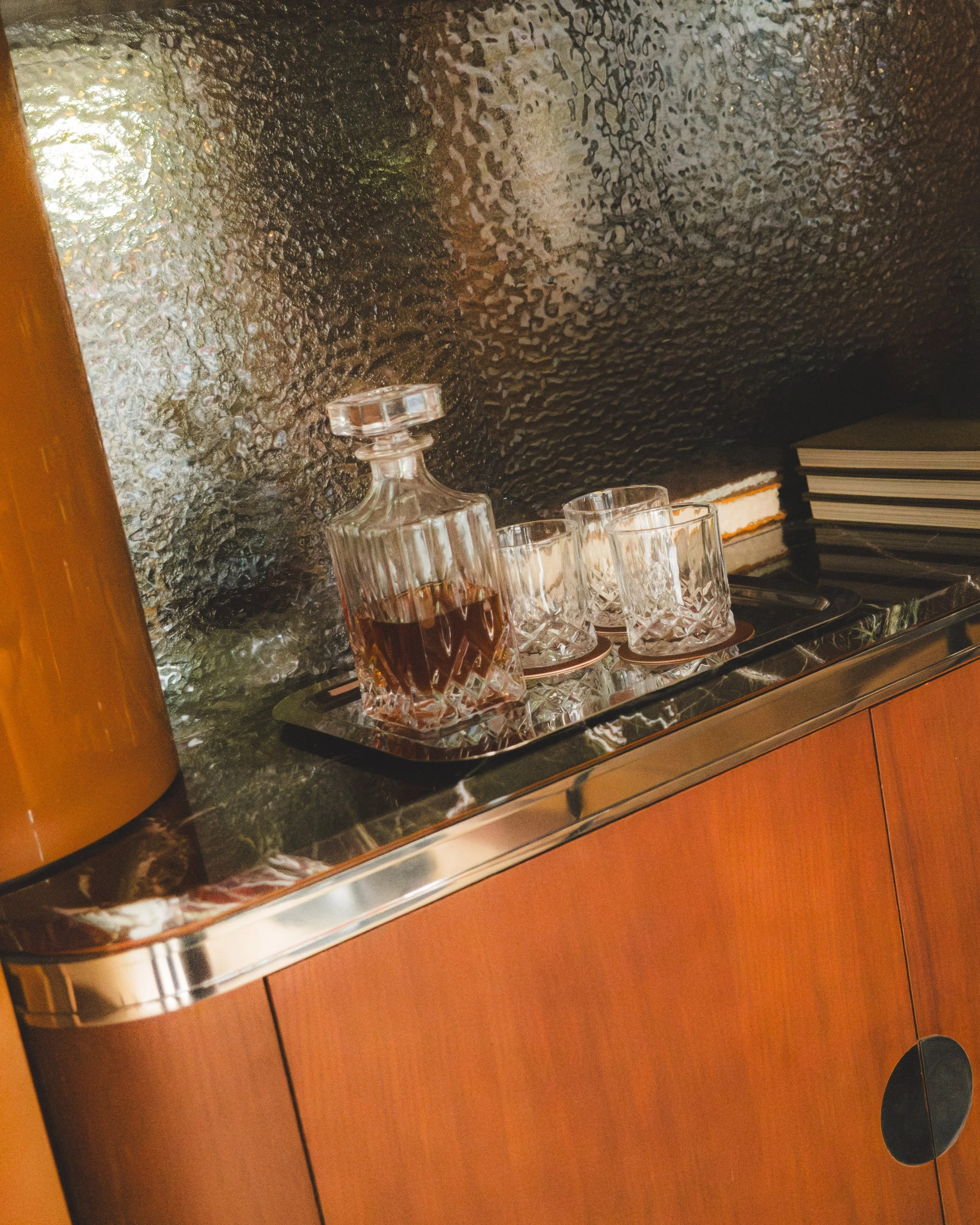 A bar or restaurant setting with a textured metallic wall in the background, a wooden counter with a glossy surface, several crystal glasses with a dark drink, and a stack of closed books or menus.