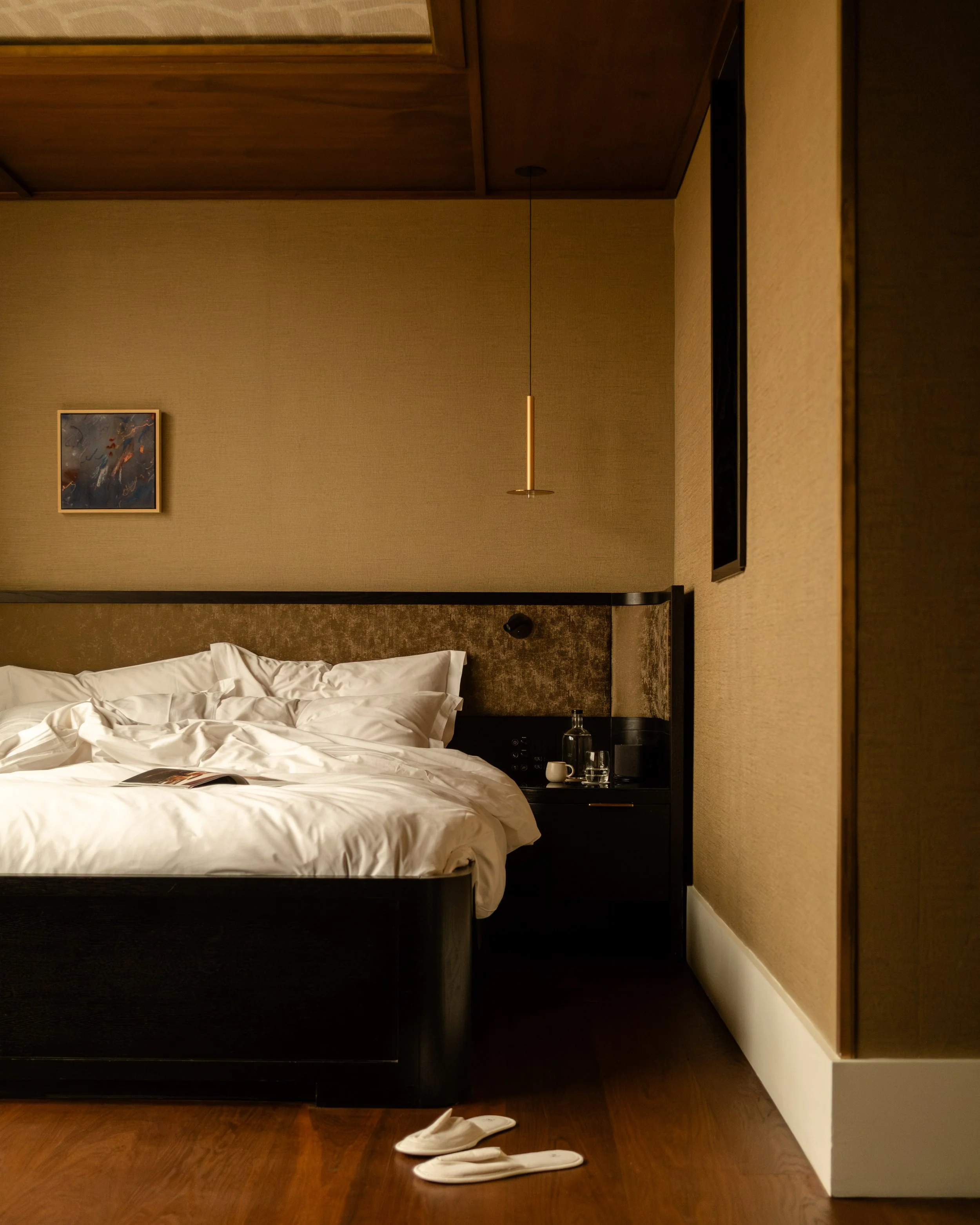A neatly made bed with white bedding and pillows, a bedside table with a bottle and glass, a small painting on the wall, and a pair of white slippers on the wooden floor. Lionel Chu - Hotel & Travel Photographer