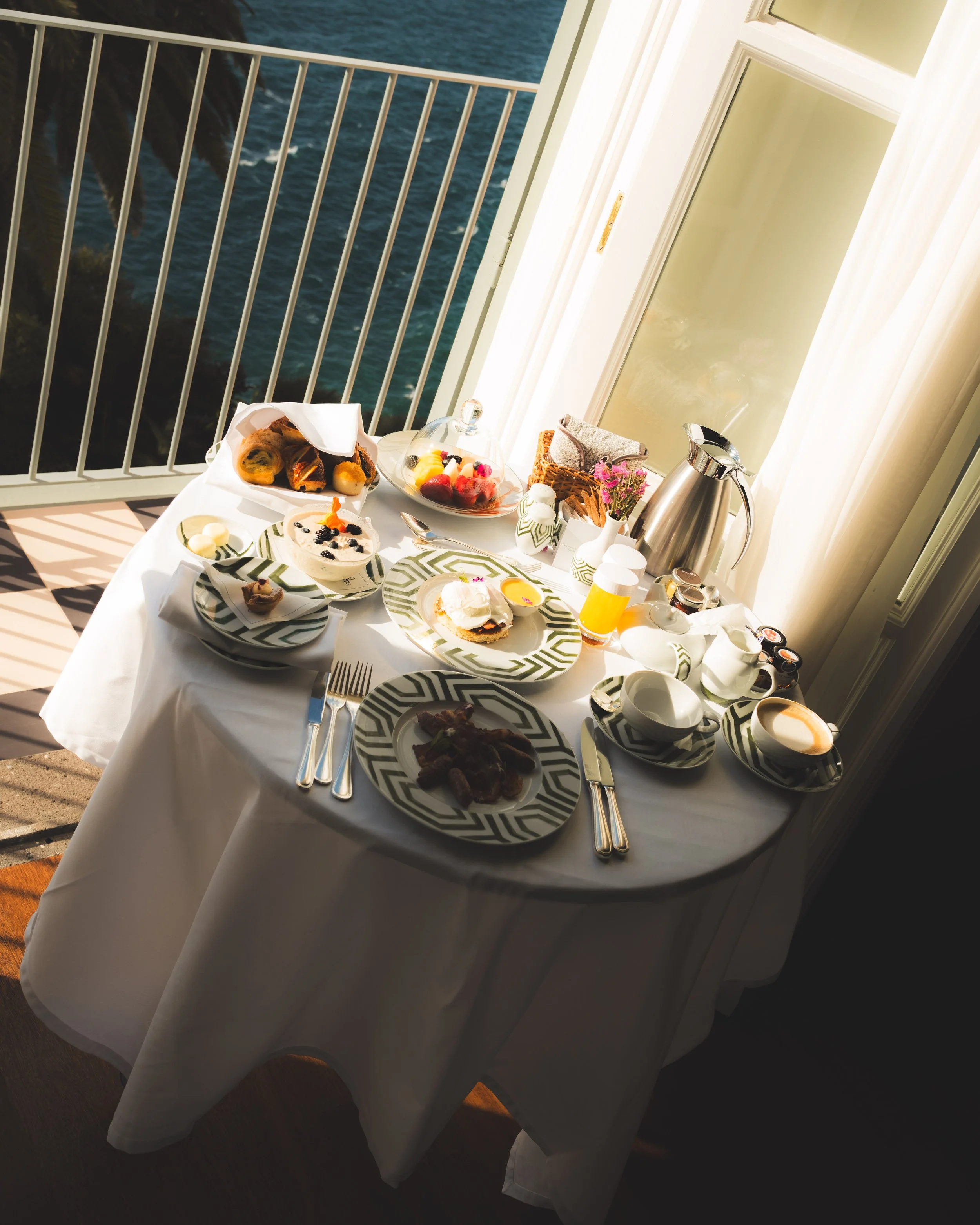 Breakfast table set on a balcony overlooking the ocean with palm trees in the background. The table has plates of assorted food, fruits, pastries, coffee, and a teapot.