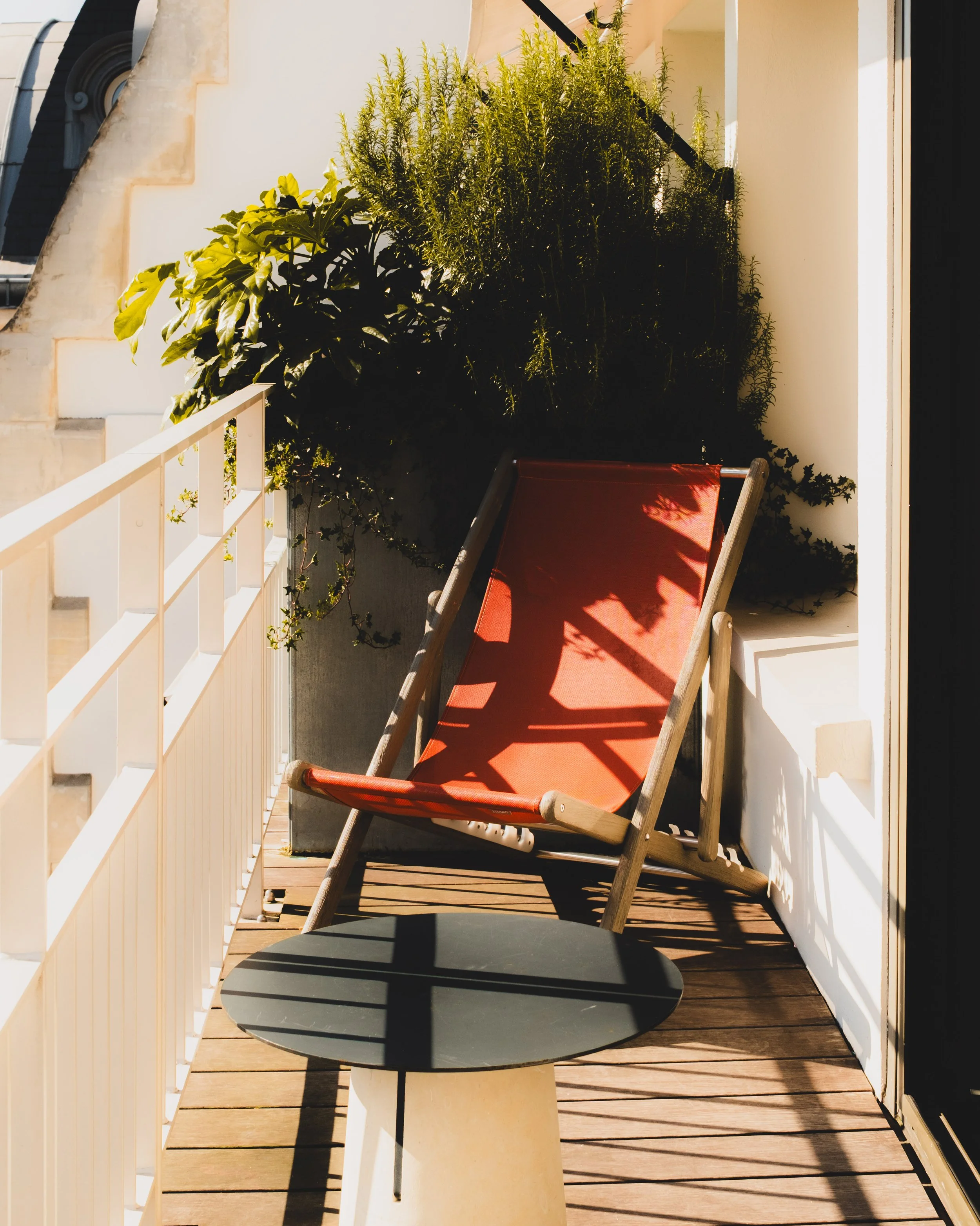 Small balcony with a black round table, a red deck chair, and green plants in the background, bathed in sunlight.