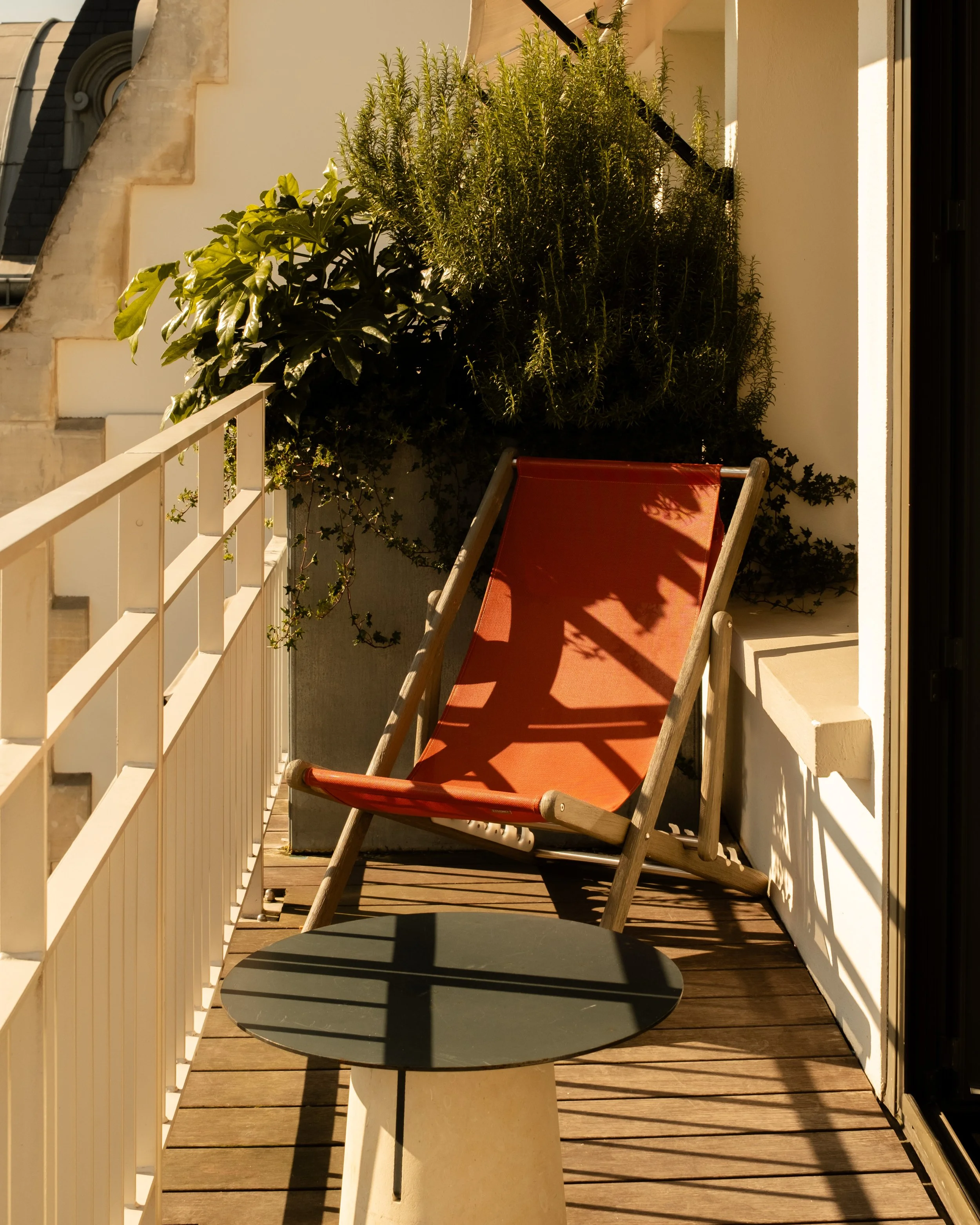 Balcony with a red deck chair, small black table, white railing, and potted green plants in sunlight. Lionel Chu - Hotel & Travel Photographer