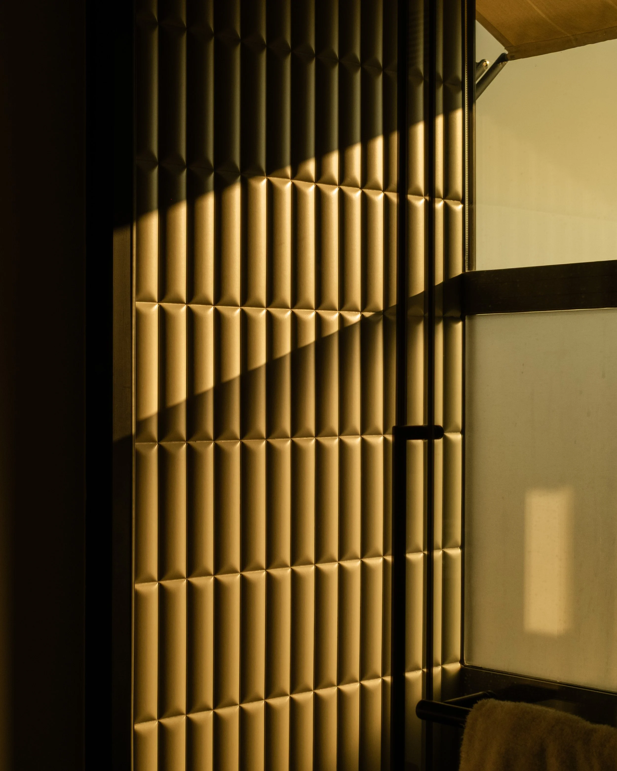 A textured, padded gold wall with a diagonal shadow, next to a glass window with a black frame and beige wall. Lionel Chu - Hotel & Travel Photographer