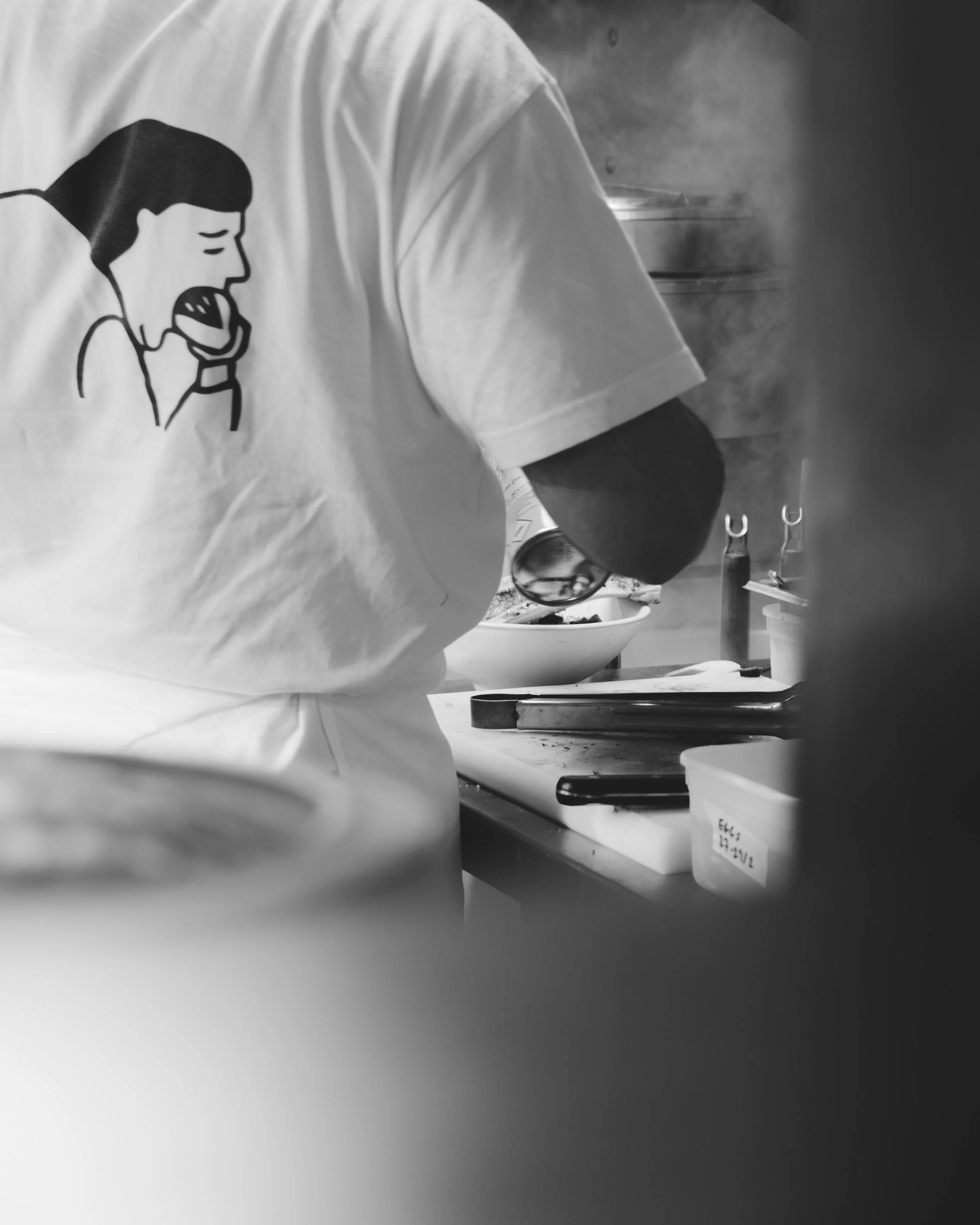 Close-up view of the back of a cook at Bao restaurant in London, wearing a t-shirt that displays the restaurant's logo (a person enjoying a bao), in the restaurant's kitchens.