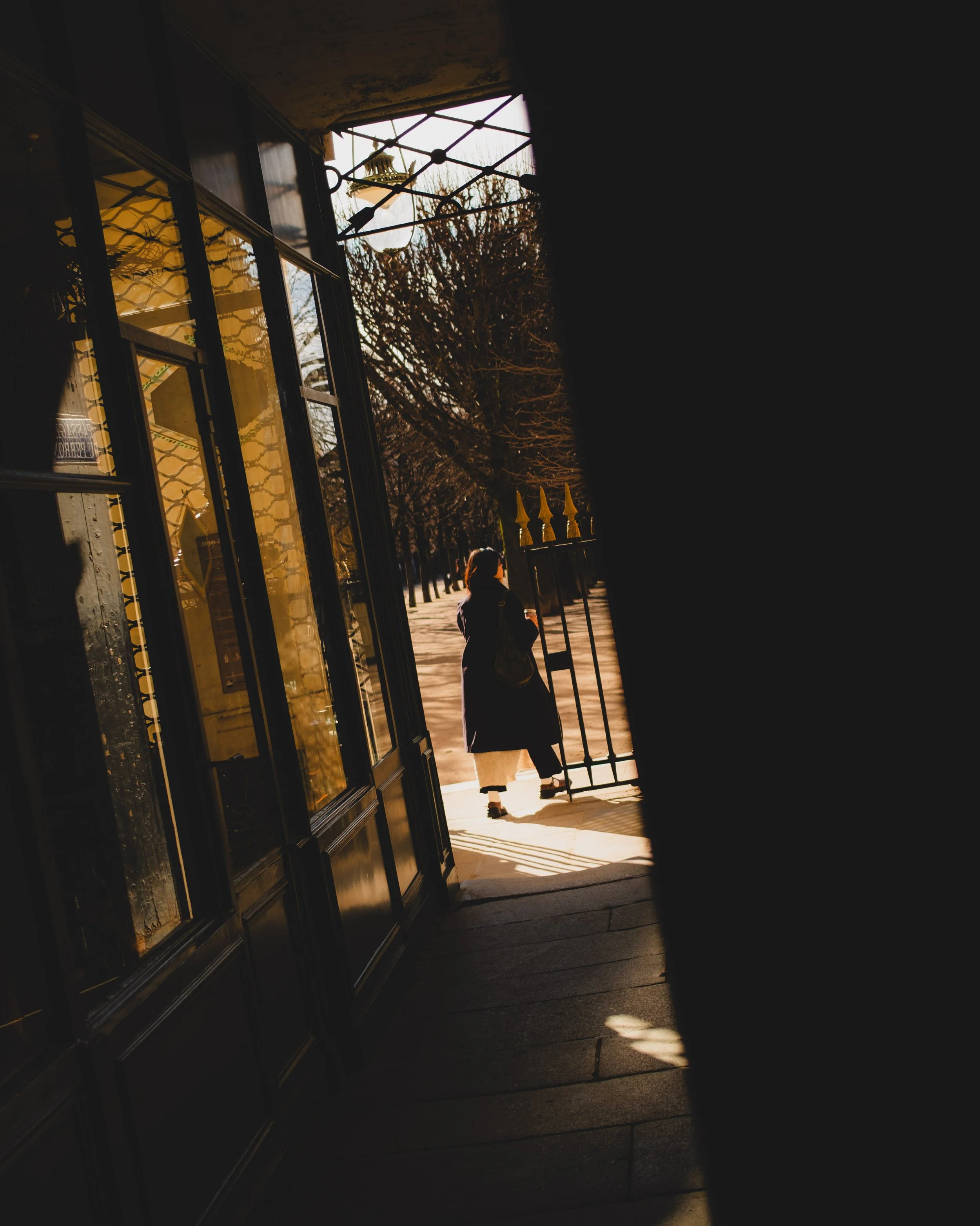 A woman stands outside a gate on a sidewalk, viewed through a partially open door, with trees and a sunny street in the background. Lionel Chu - Hotel & Travel Photographer