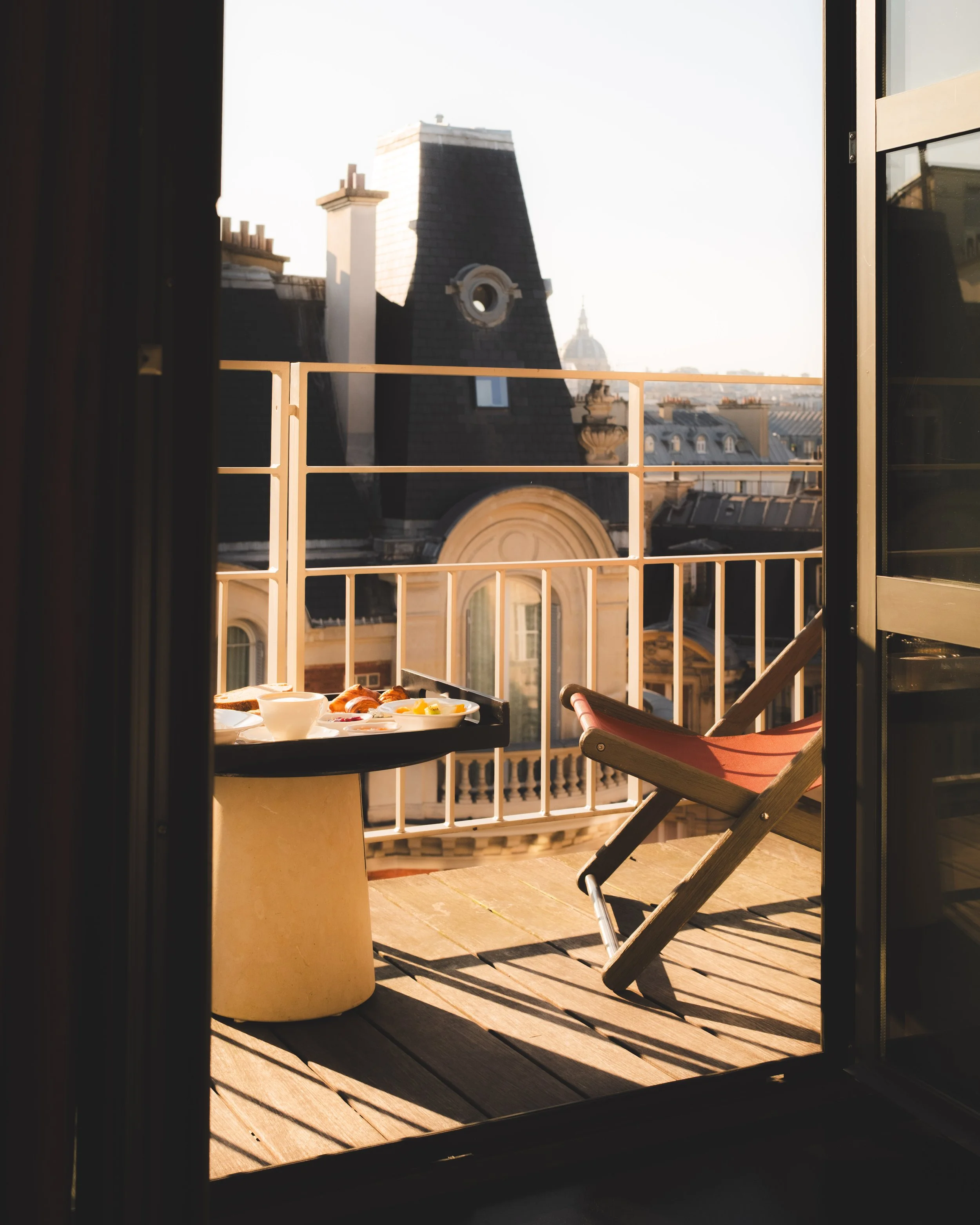 View through a doorway showing a balcony with a table set for breakfast, a folding chair, and Parisian rooftops with domed buildings in the distance at sunrise.