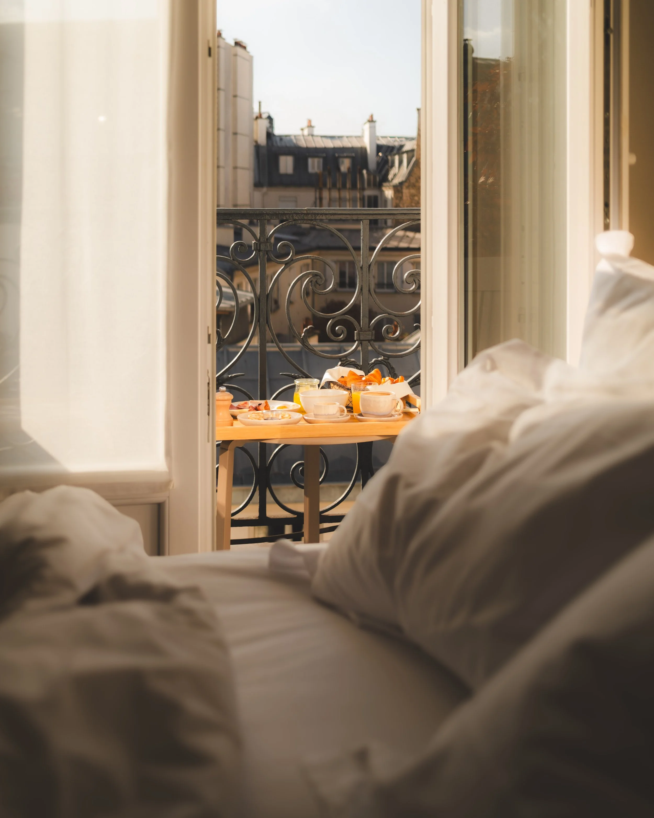 A doorway open to a balcony with a view of neighboring buildings and rooftops, as seen from a bed with white sheets.