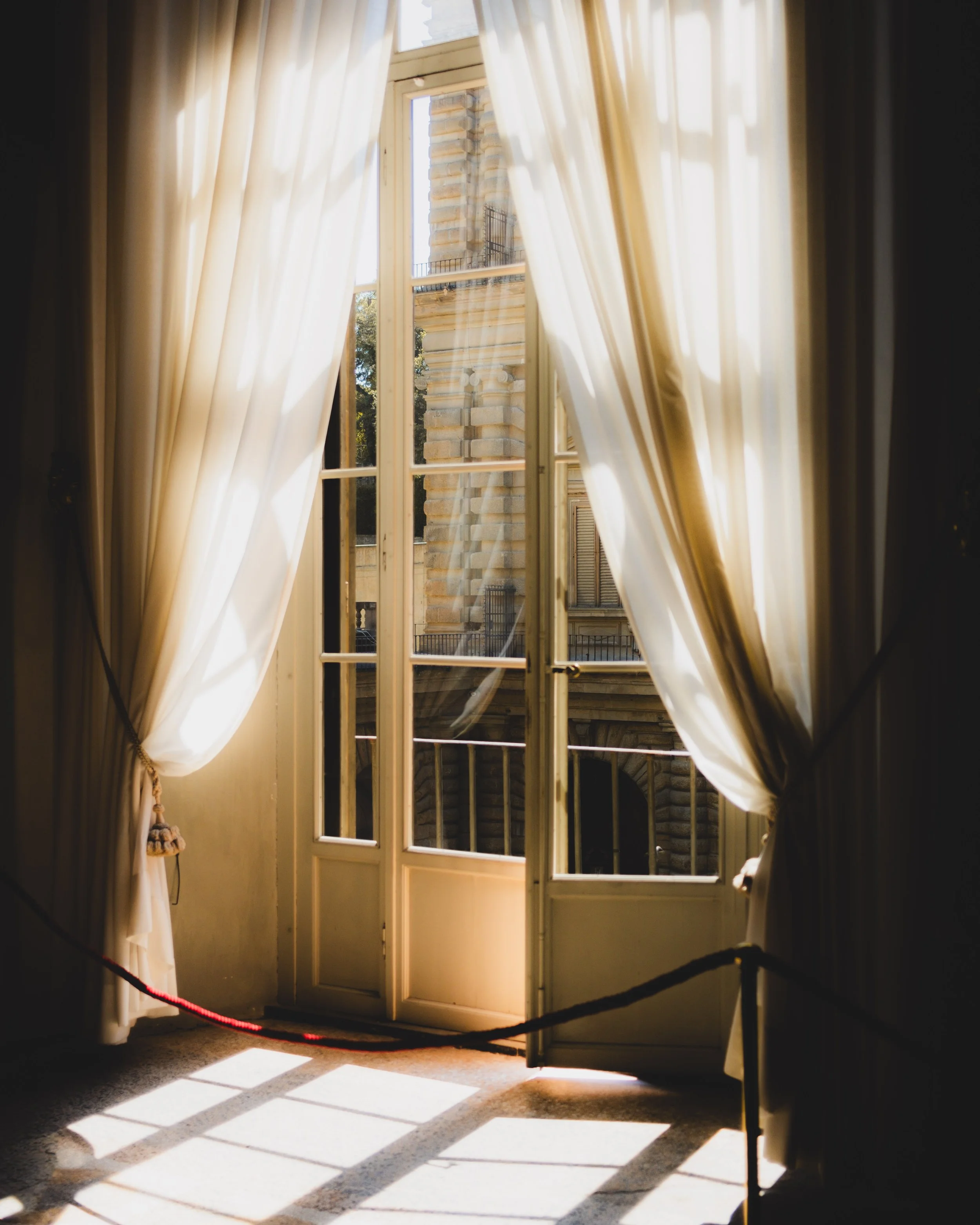 Open glass doors with cream curtains and sunlight casting shadows inside a room, with a building visible outside. Lionel Chu - Hotel & Travel Photographer