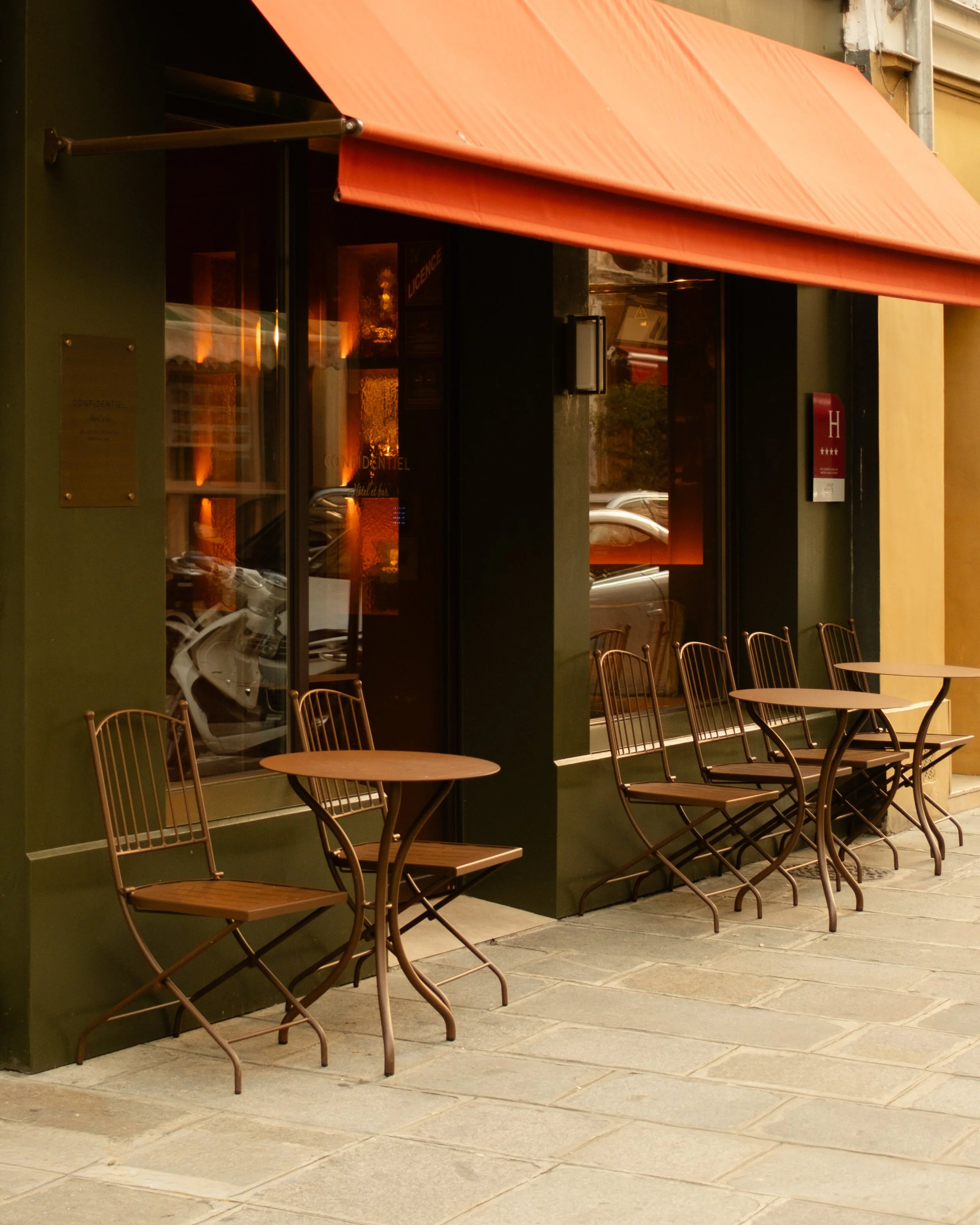 Empty outdoor seating with four wooden chairs and four round tables outside an hotel with a large orange awning Lionel Chu - Hotel & Travel Photography