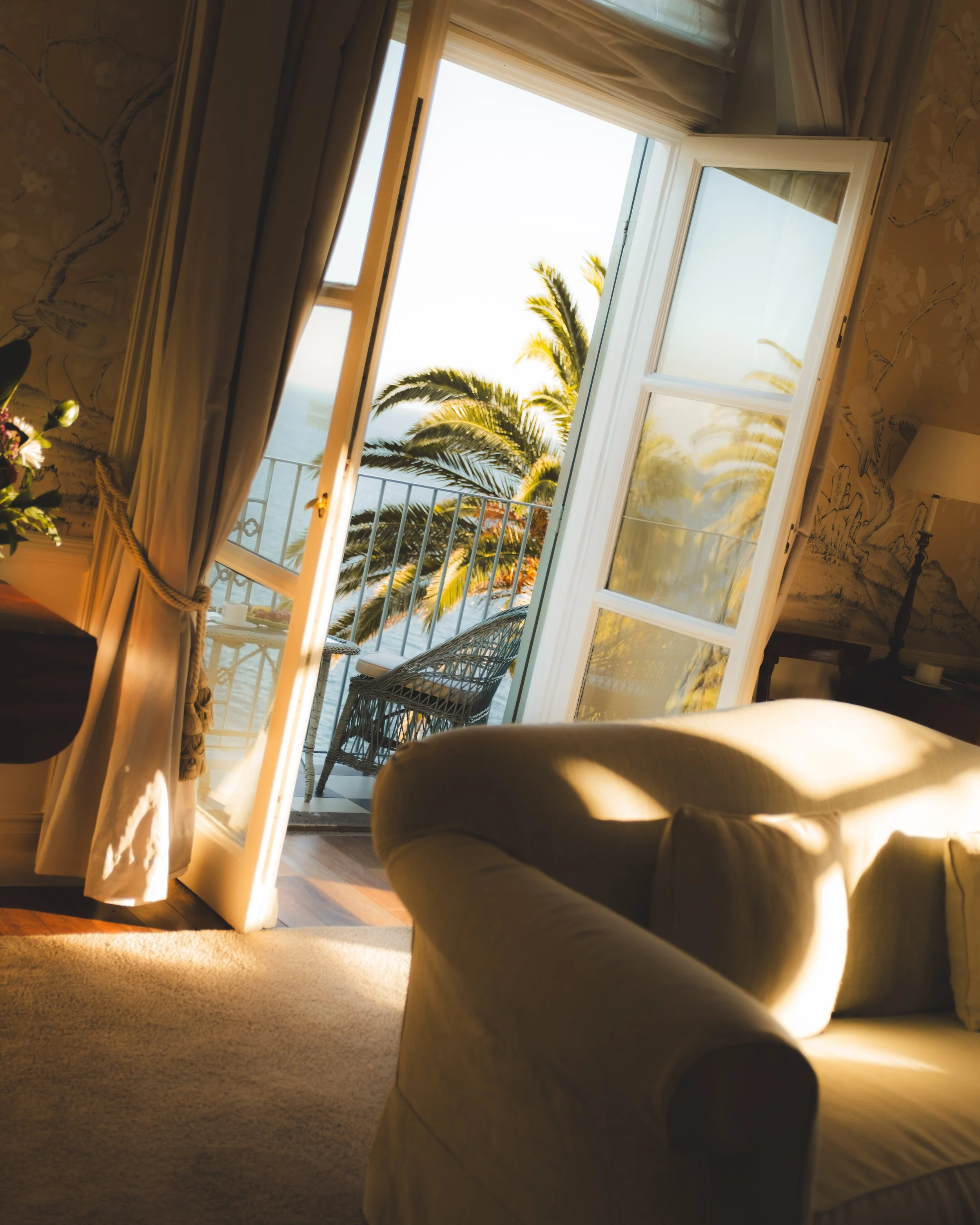 A view from inside a room looking out through open French doors to a balcony with a railing, tropical palm trees, and the ocean in the background during sunset or sunrise.