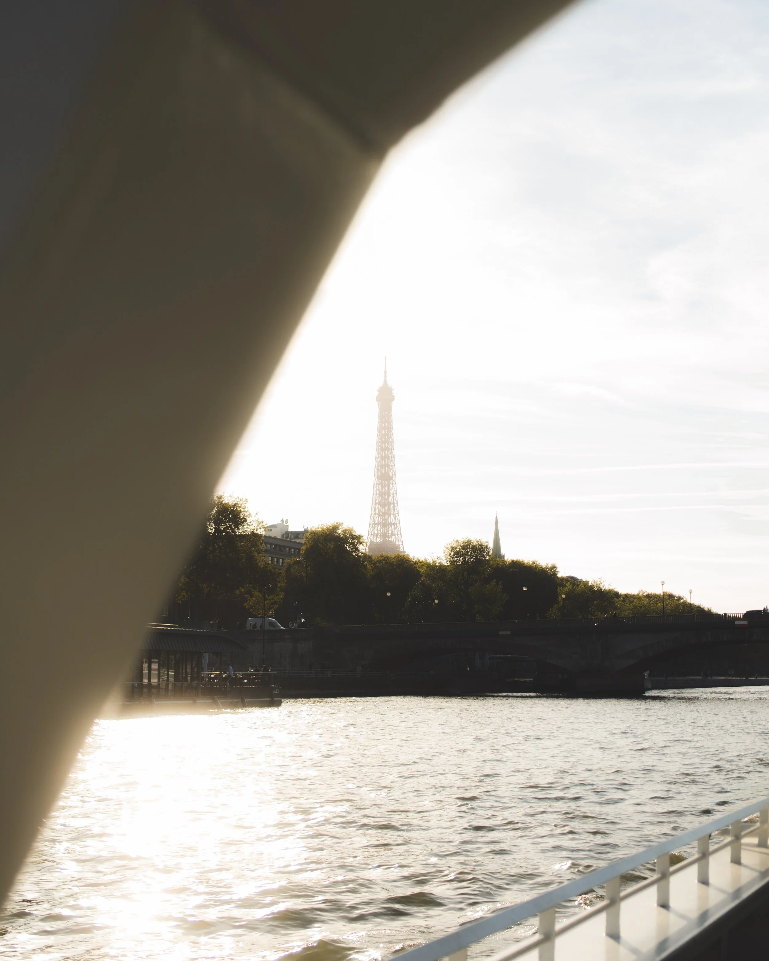 View of the Eiffel Tower in Paris, France, seen through a gap in a ship's structure, with water and a bridge in the foreground and a partly cloudy sky.