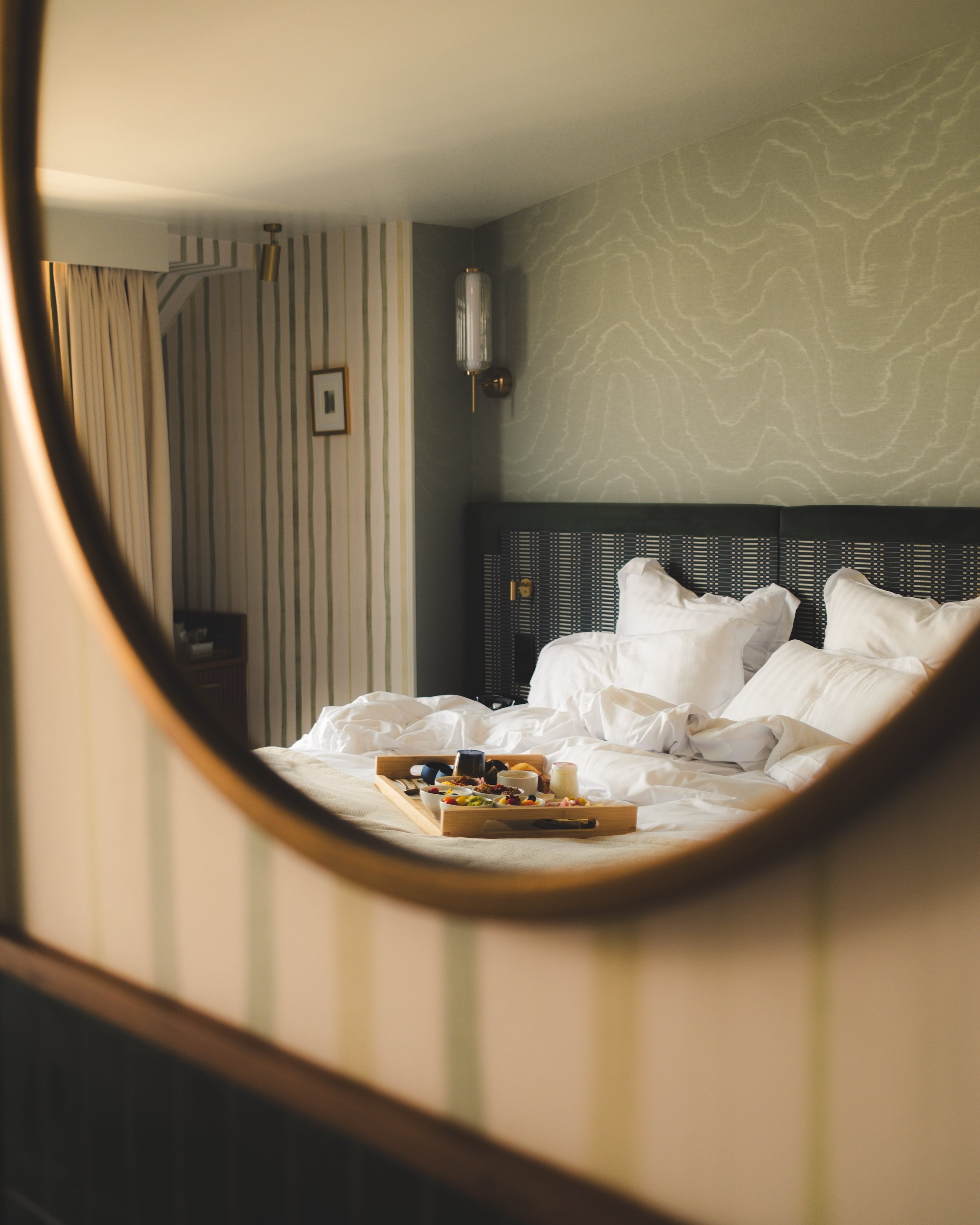 A bedroom seen through a circular mirror, showing a bed with white linens, a tray with food on the bed, and a wall with striped wallpaper and decorative lighting.