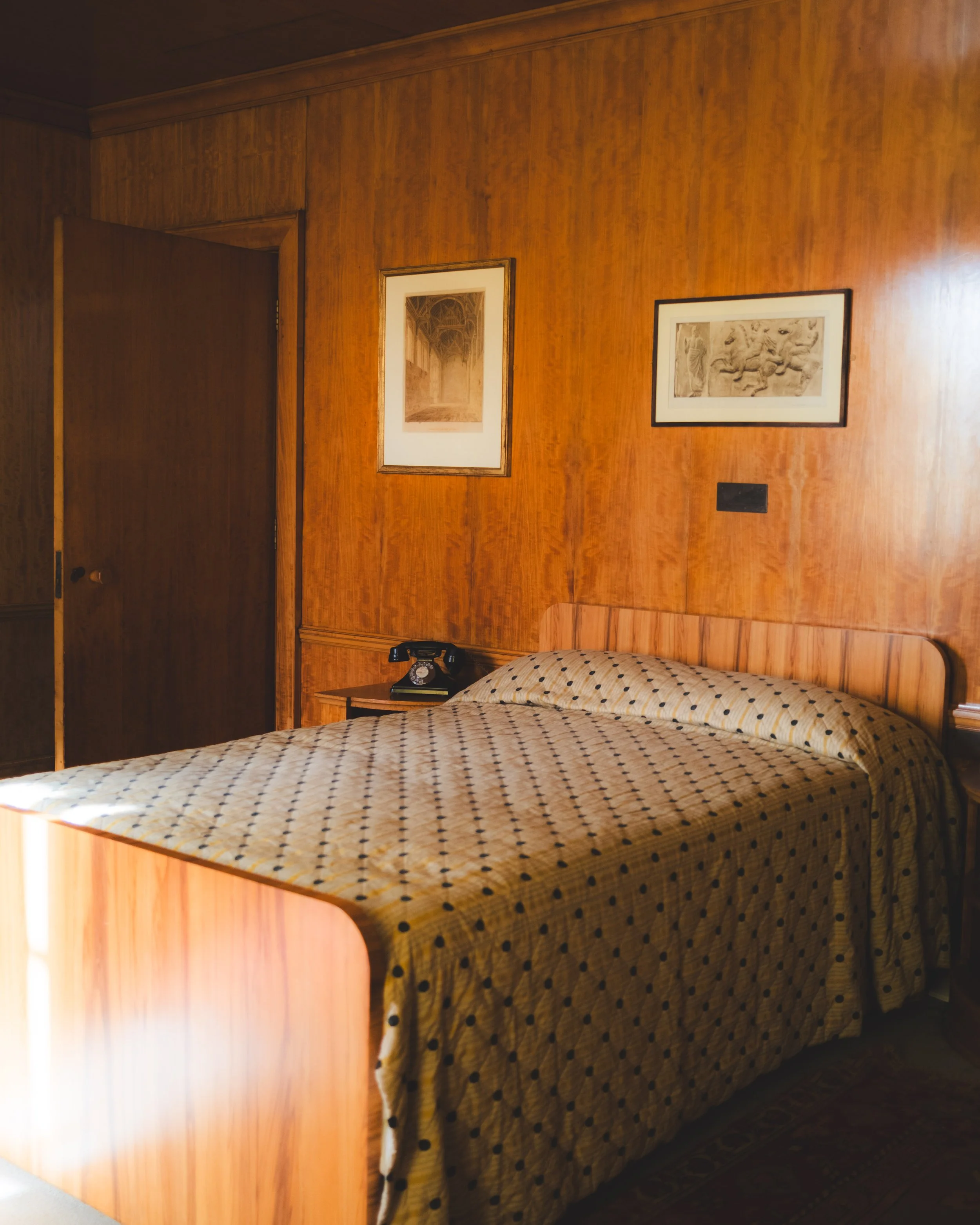 A bedroom with wood-paneled walls, a bed with a yellow and black polka dot blanket, a bedside table with a black rotary phone, and framed artwork on the wall.