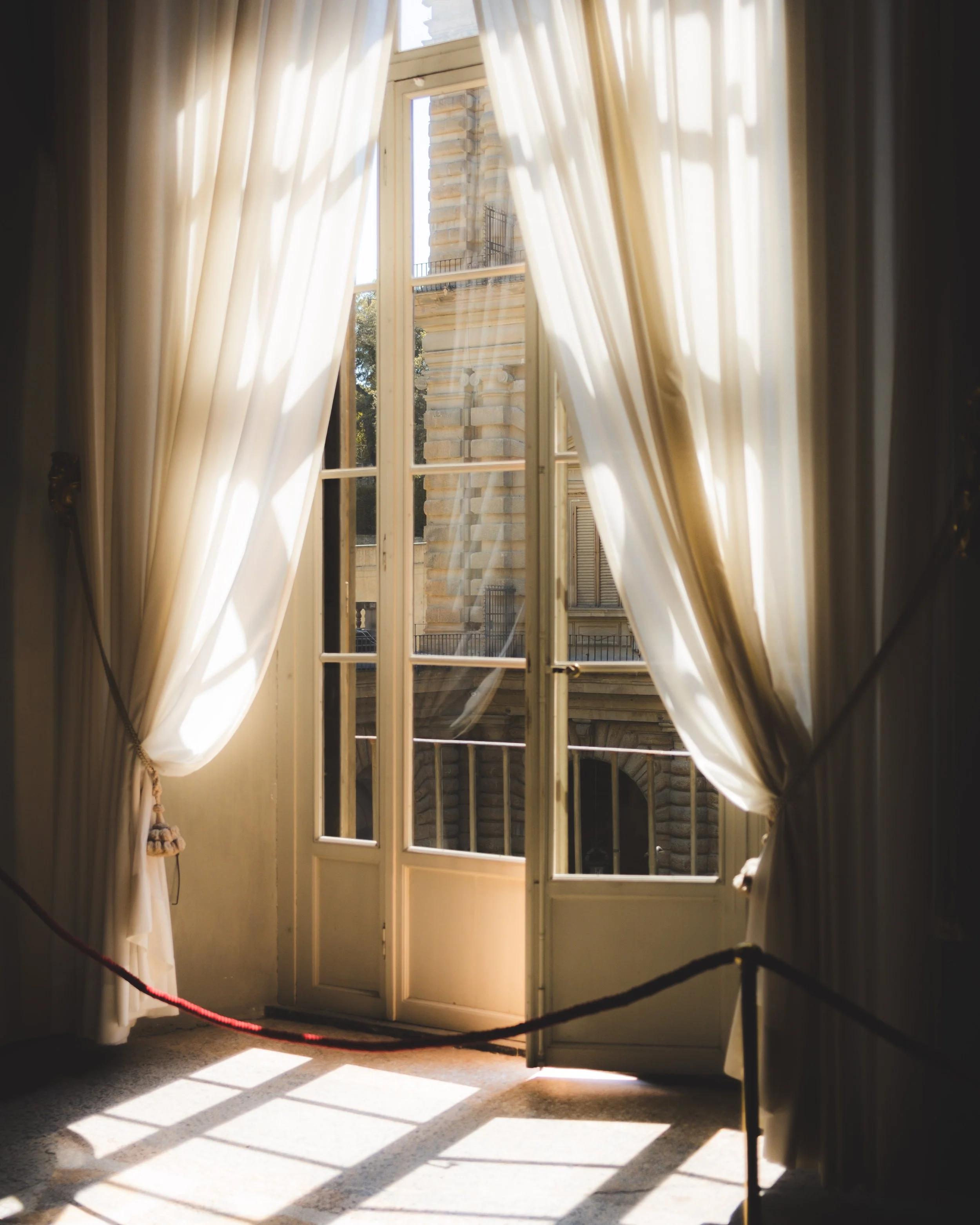 Open glass windows doors with cream curtains and sunlight casting shadows inside a room, with a building visible outside. Lionel Chu - Hotel & Travel Photographer