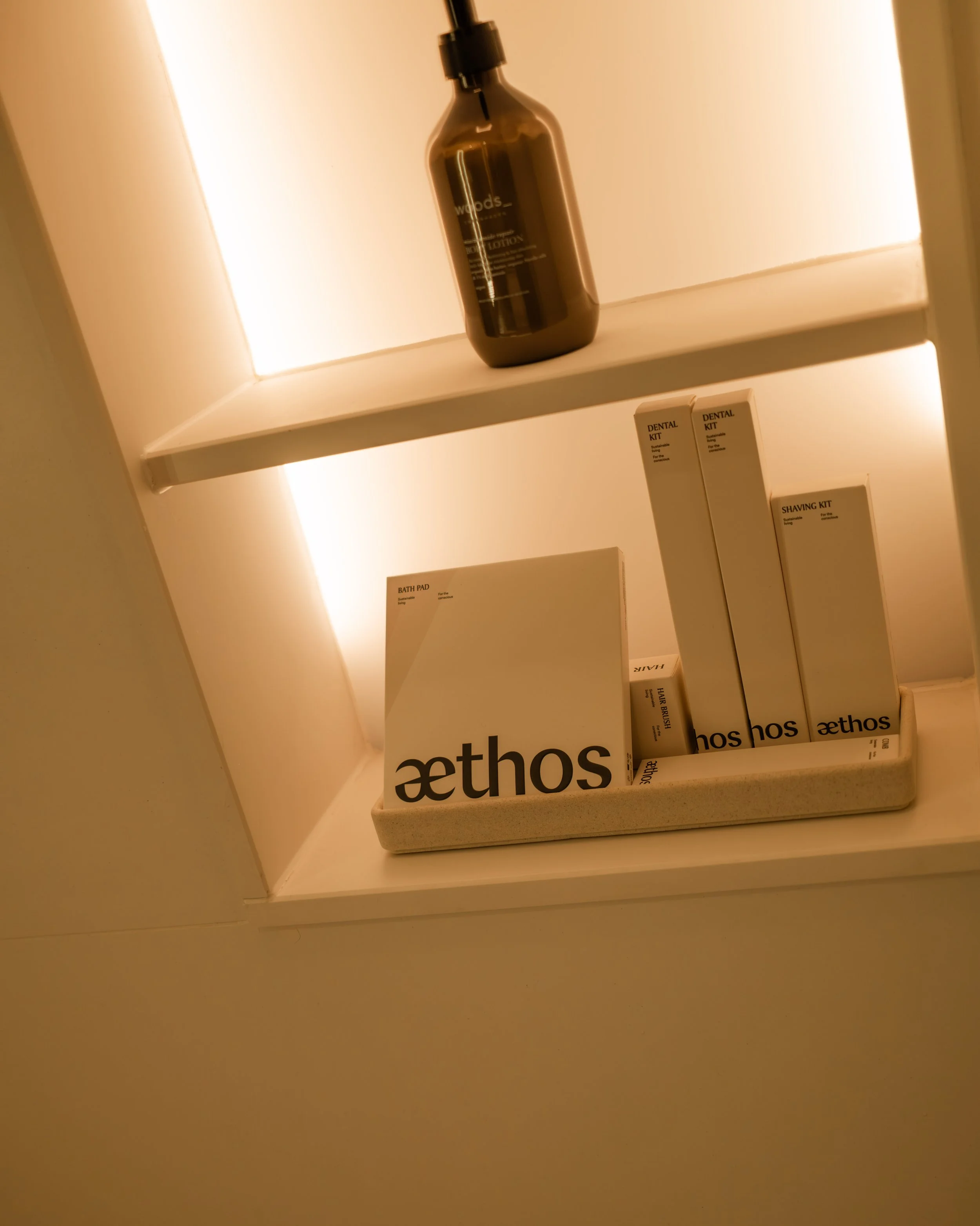 Shelves with Aethos-branded bathroom products, including bath pad, dental kits, and shaving kit, with a brown bottle on the top shelf. Lionel Chu - Hotel & Travel Photographer