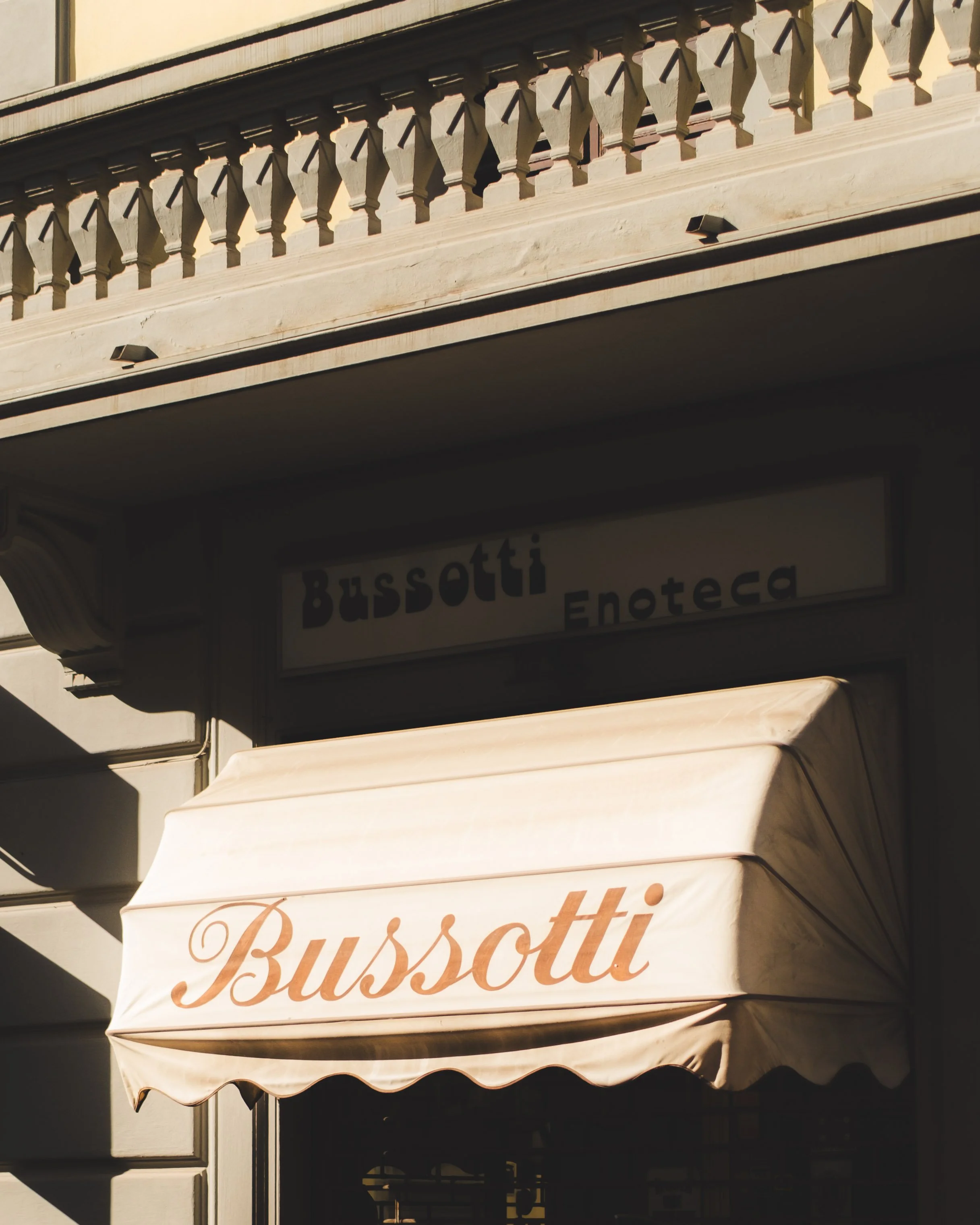 Storefront of Bussotti Enoteca with a beige awning displaying the name 'Bussotti' in brown cursive lettering, set outside a building with a decorative balcony above. Lionel Chu - Hotel & Travel Photographer