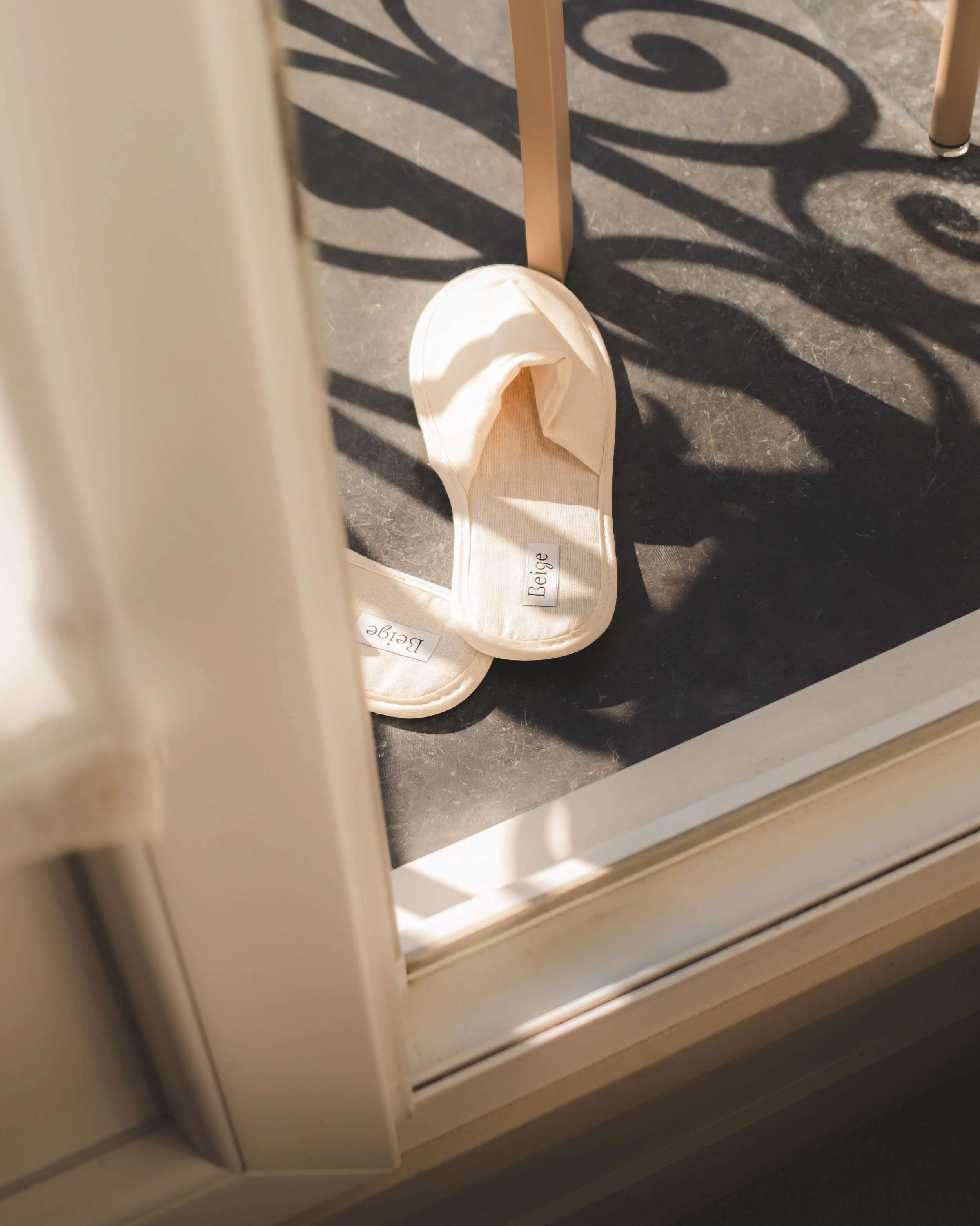 A pair of beige slippers with labels that say 'Beige' on the inside, on a dark gray floor near a window with white framing and a decorative black iron railing casting shadows on the ground.