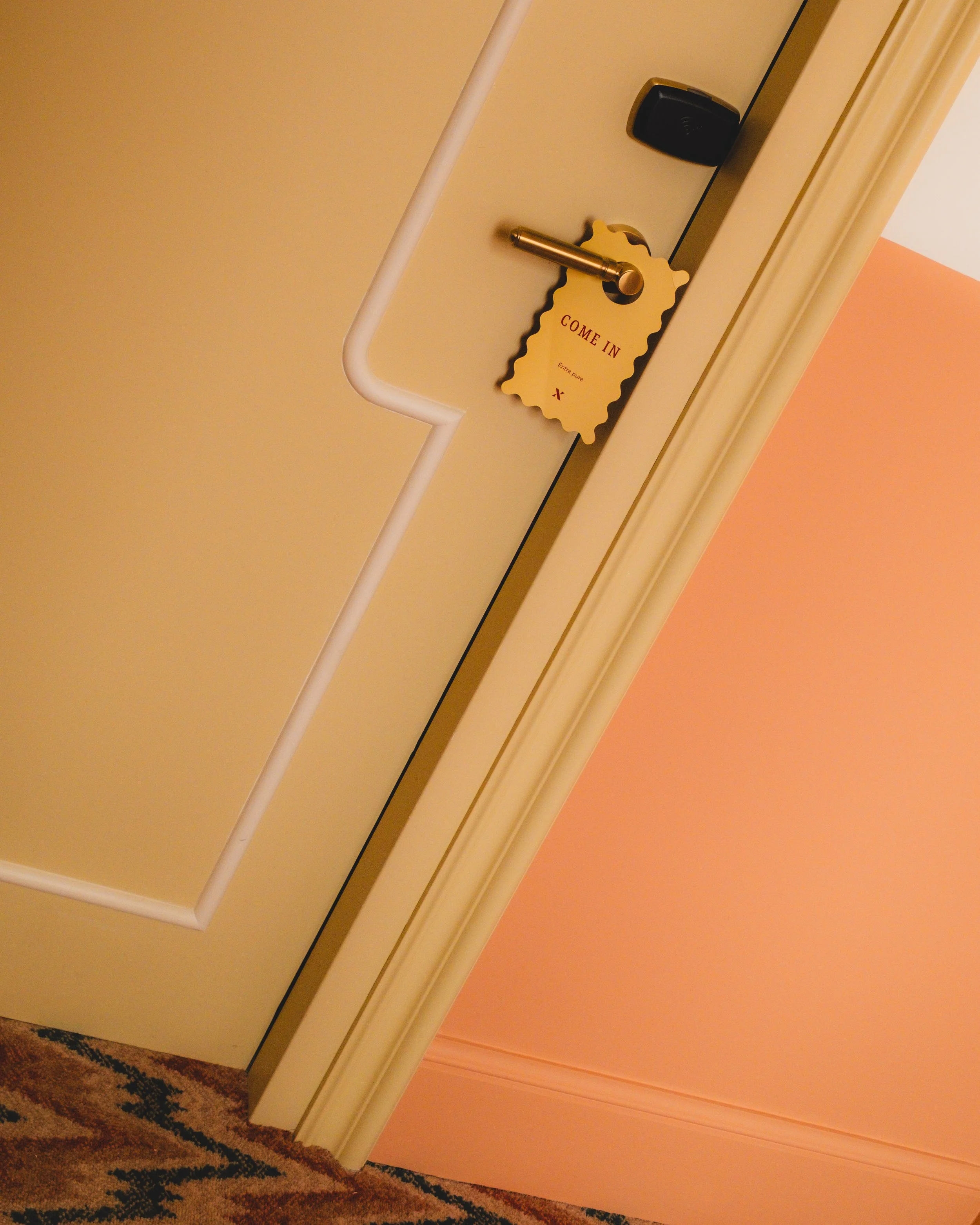 A hotel door with a golden handle and a door sign that reads 'Come In.' The door has a white safety bar, and the door frame is painted cream. The wall next to the door is painted in a salmon pink color, and there is a dark-colored coat hook above the sign. The floor has a patterned carpet with earthy tones.