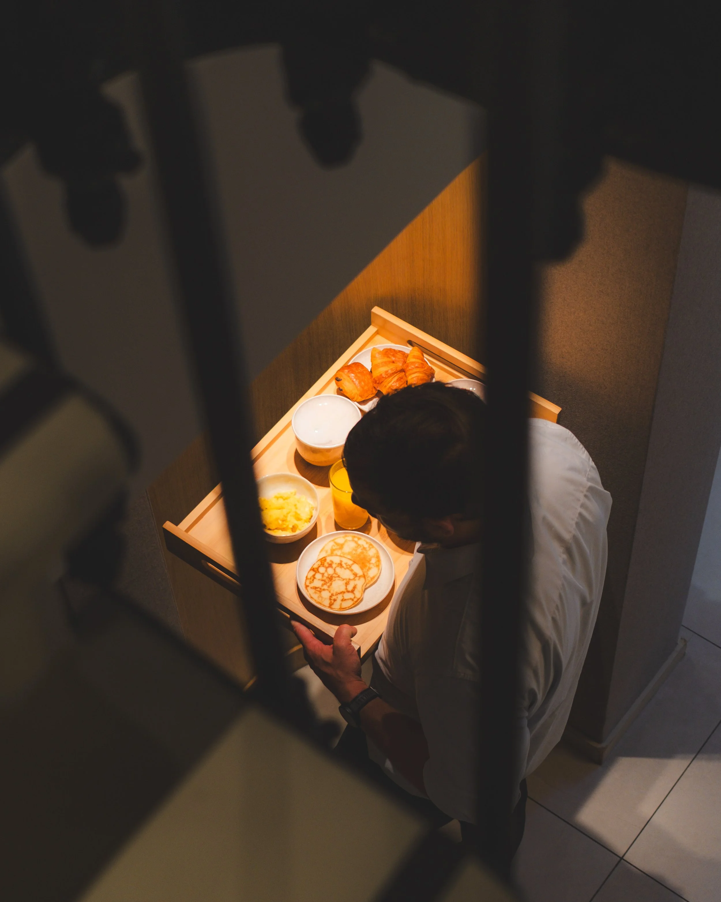 A person wearing a white shirt is standing in front of a small table filled with breakfast items such as pancakes, croissants, scrambled eggs, and cups of orange juice. The photo is taken through a fence or grill, partially obscuring the view.