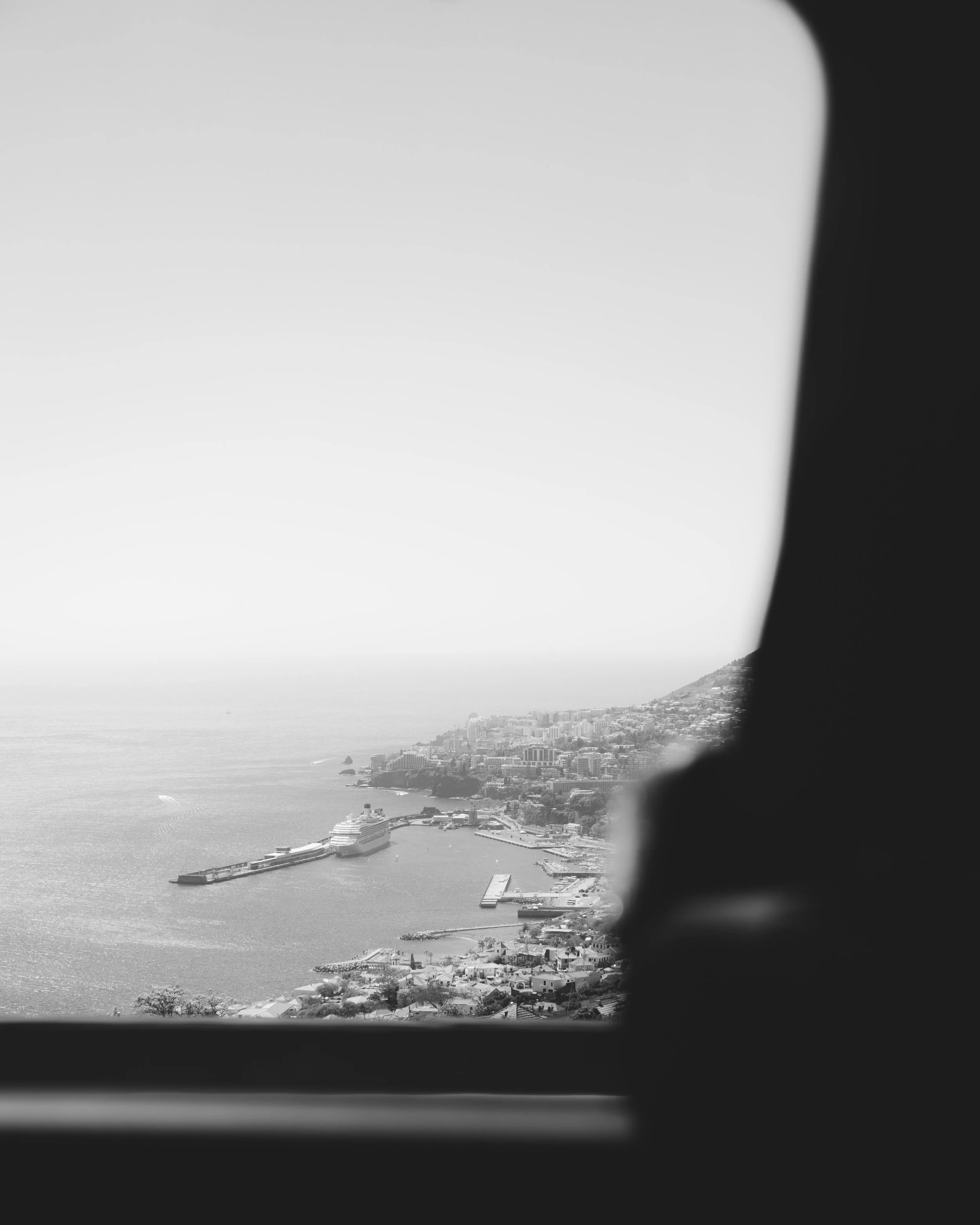 An aerial view from a car window of Funchal in Madeira with a cruise ship docked at the pier, surrounded by buildings, hills, and the ocean under a hazy sky.