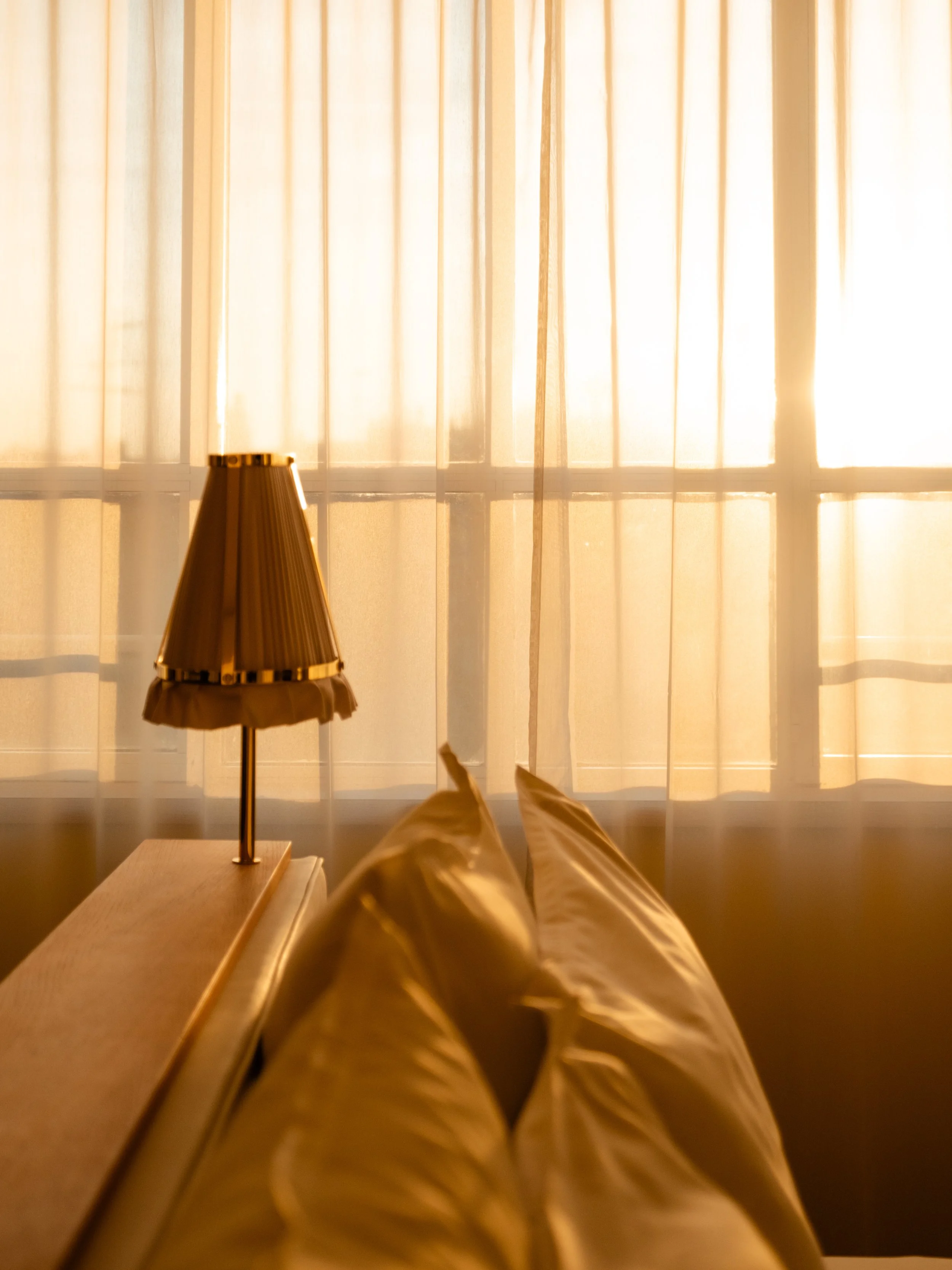 A person lying on a bed looking at sunlight coming through closed curtains, with a table lamp near them.