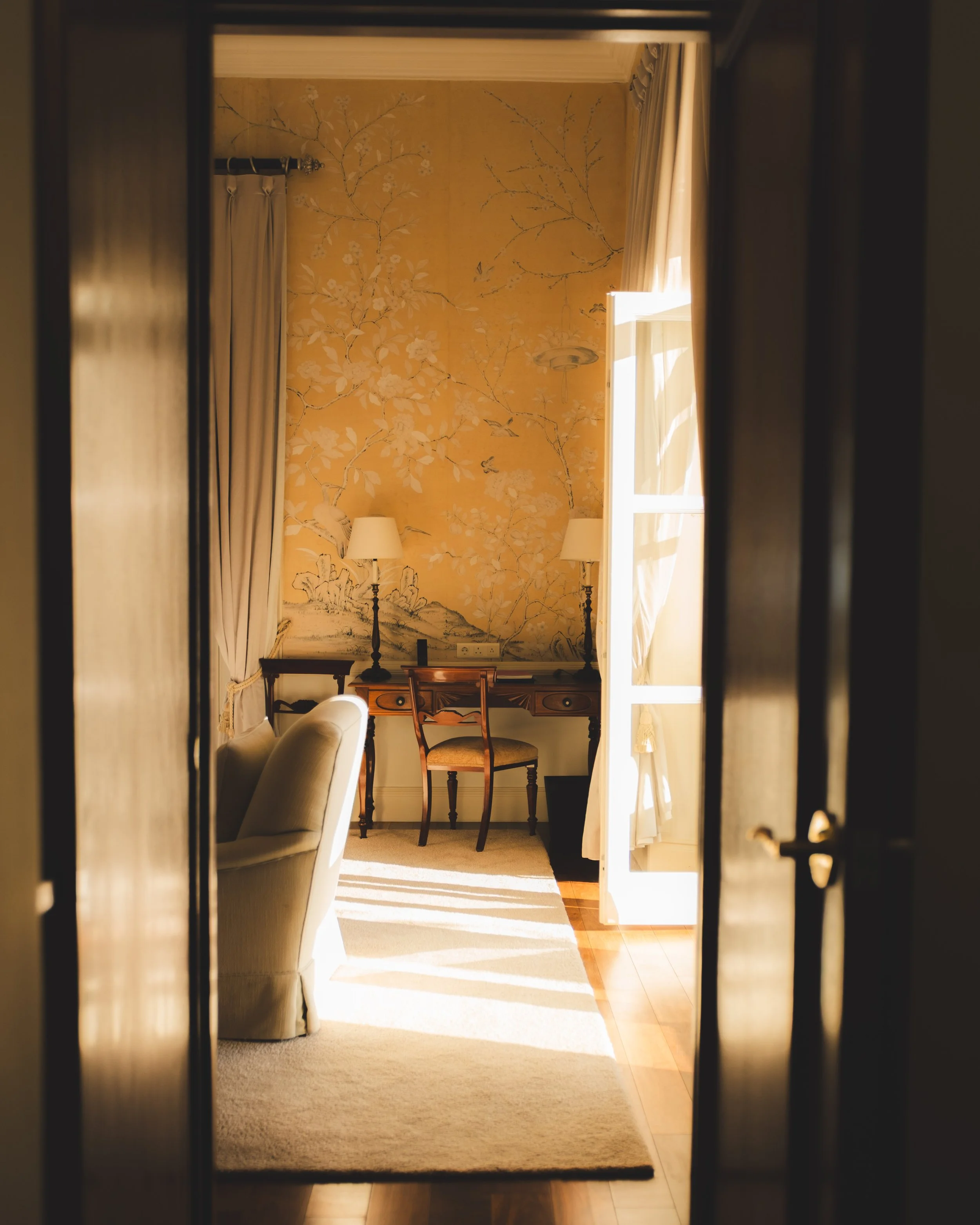 Looking through a doorway into a living room with a yellow floral wallpaper, a window with white curtains, and furniture including a sofa, chairs, and lamps.