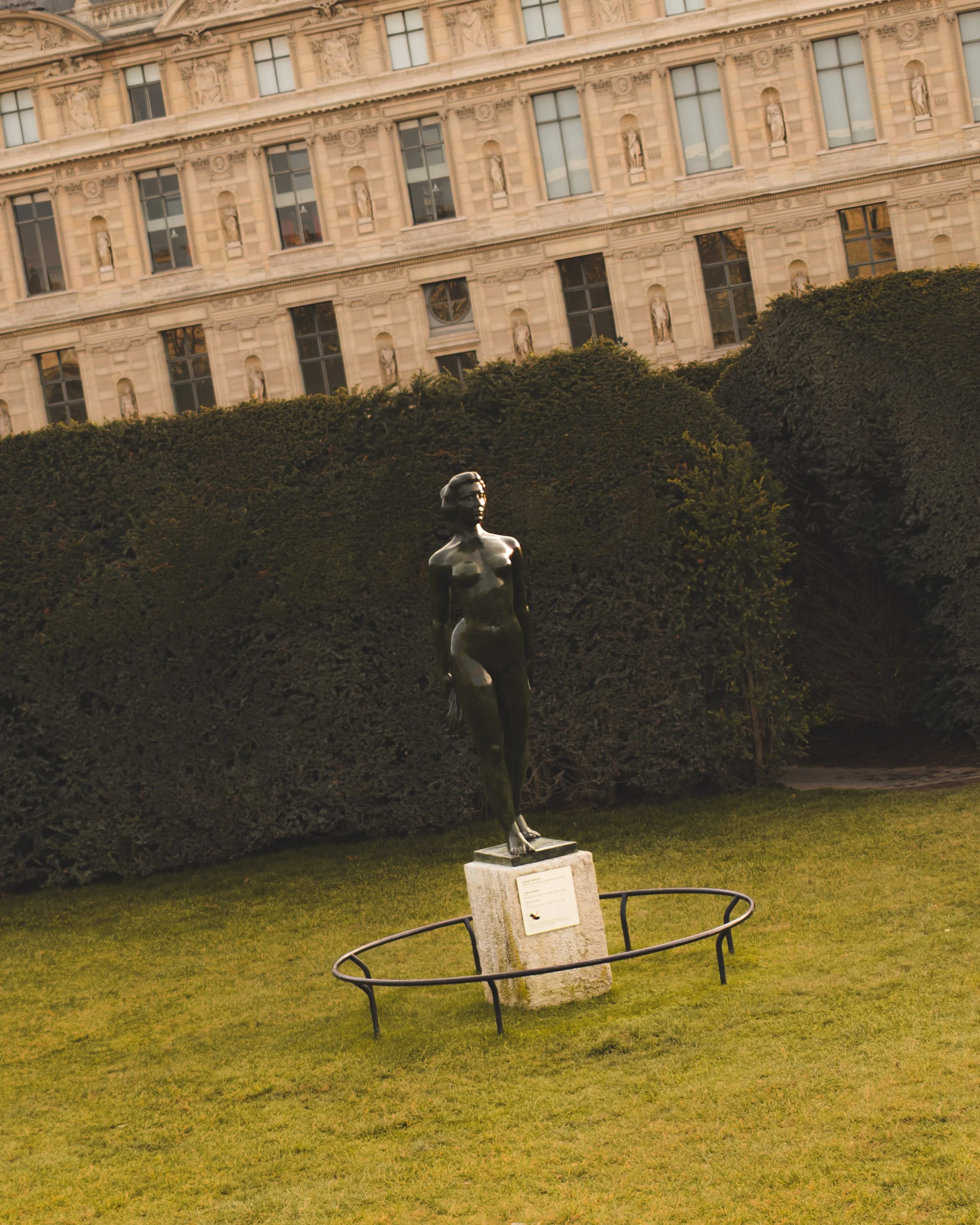 A metallic statue of a woman stands in front of a historic building with large windows and decorative statues. The statue is surrounded by a circular metal fence and is positioned on a stone pedestal with an inscription plaque.